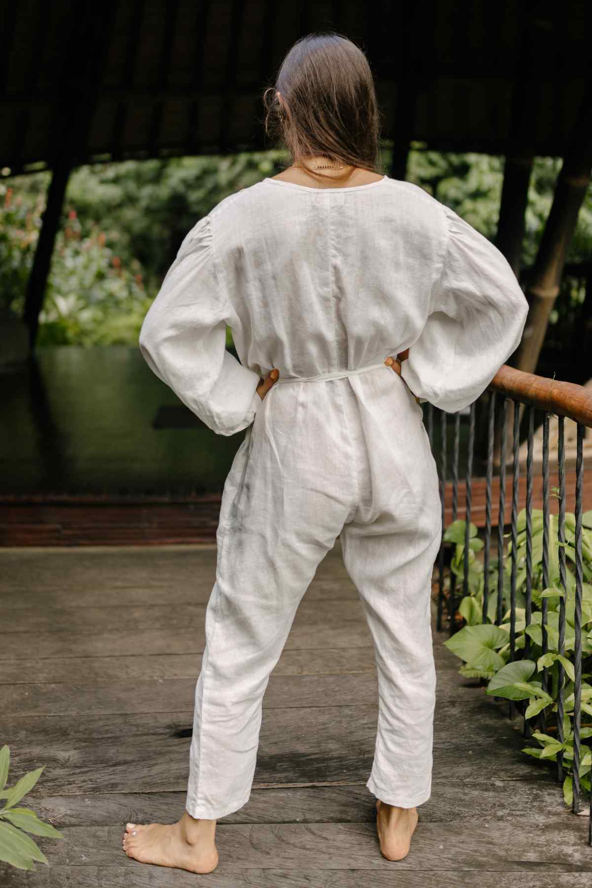 A person with long hair sits cross-legged on a mat, holding a small item with eyes closed, appearing to meditate. They are wearing a light-colored Linen Long Sleeve Dora Playsuit from Myrah Penaloza, featuring unique detailing and an adjustable waistline, in a sunlit wooden structure with a peaceful ambiance.