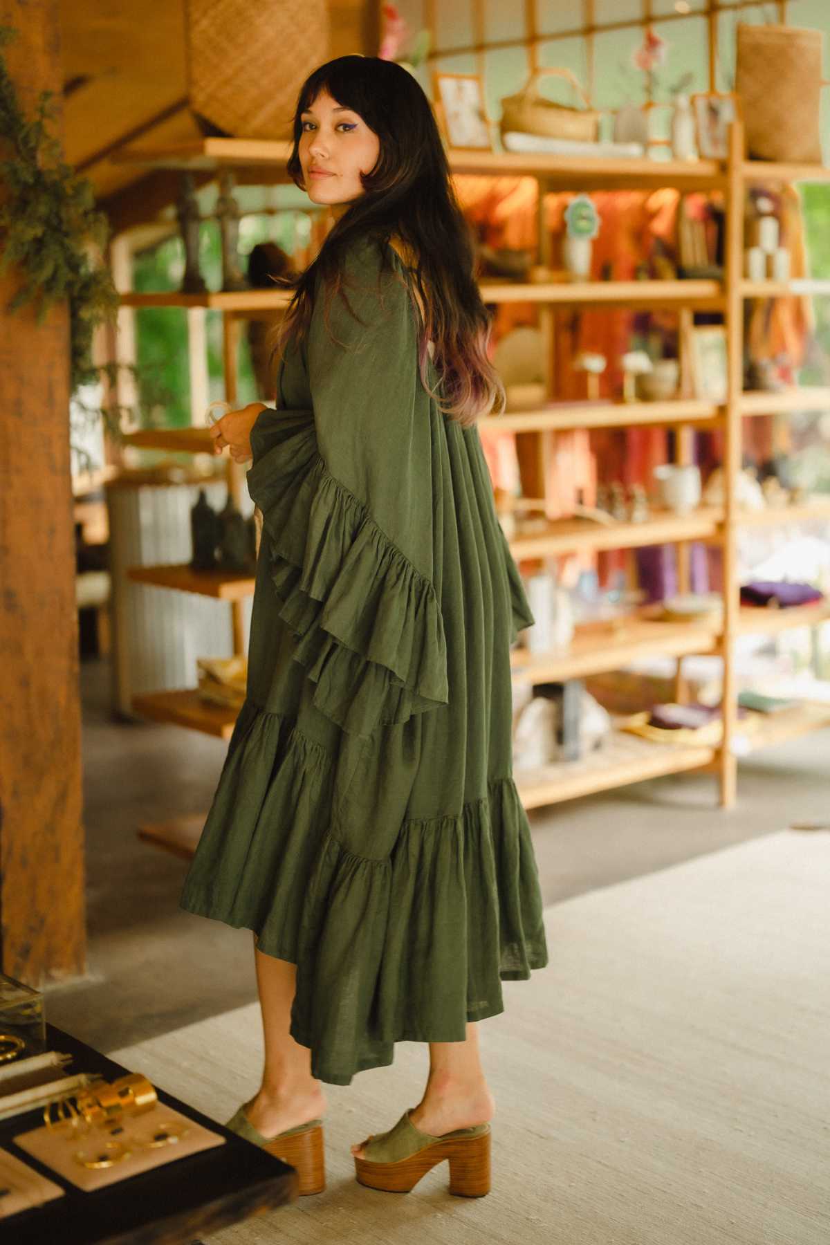 A person in a green, ruffled Brida Linen Gown Long by Myrah Penaloza and platform shoes stands in a store with wooden shelves of various items. The cozy ambiance is softly lit, while carbon neutral shipping promises an eco-friendly shopping experience.