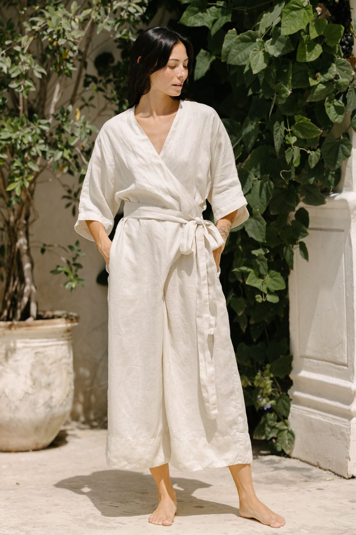 A person with long dark hair stands barefoot outdoors on a sunny day. They are wearing the Rey Linen Playsuit by Myrah Penaloza, which is light-colored and loose-fitting with long sleeves and an adjustable front wrap tied at the waist. They are standing in front of a large potted plant with lush green foliage.