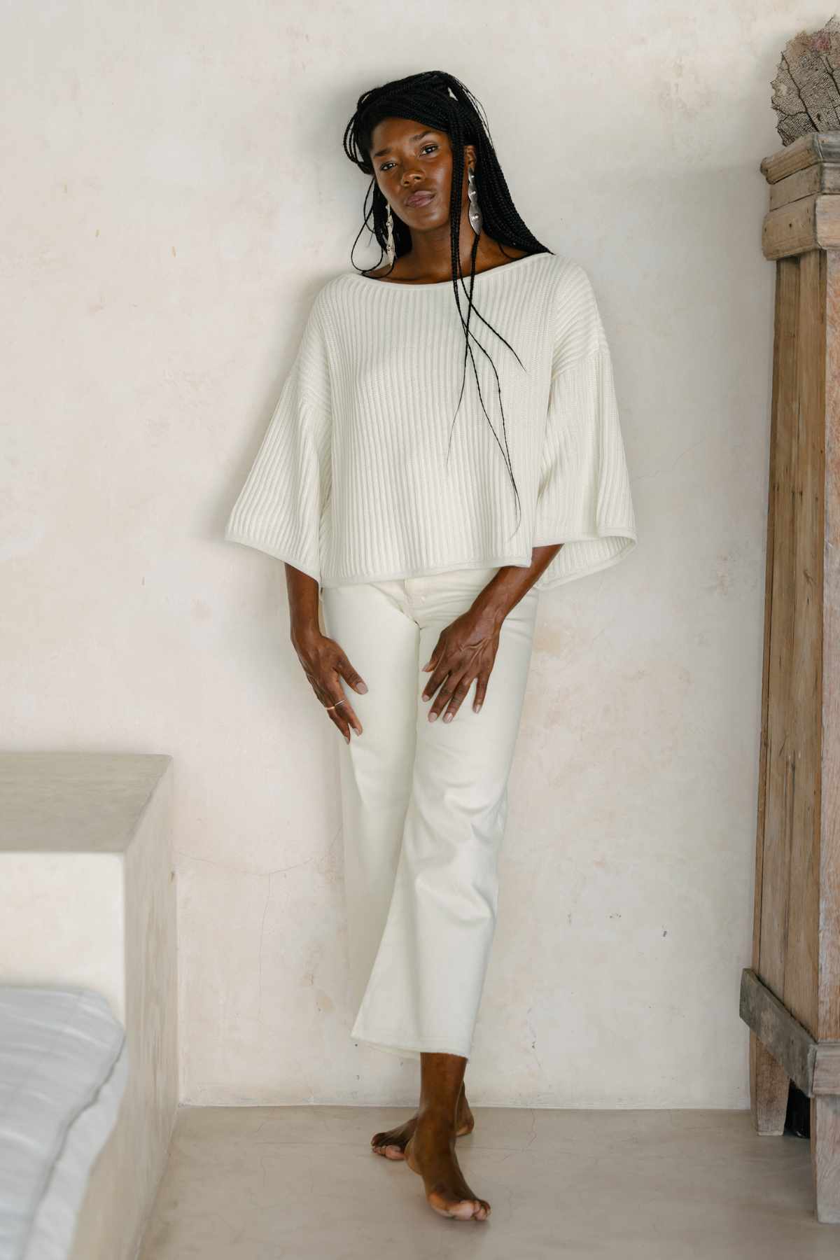 A person stands barefoot indoors against a light-colored wall, wearing an oversized Spirit Top Sweater Knit by Myrah Penaloza in off-white and white pants. Their long hair is styled in braids, and they have a relaxed, composed demeanor with hands gently touching their thigh and hip.