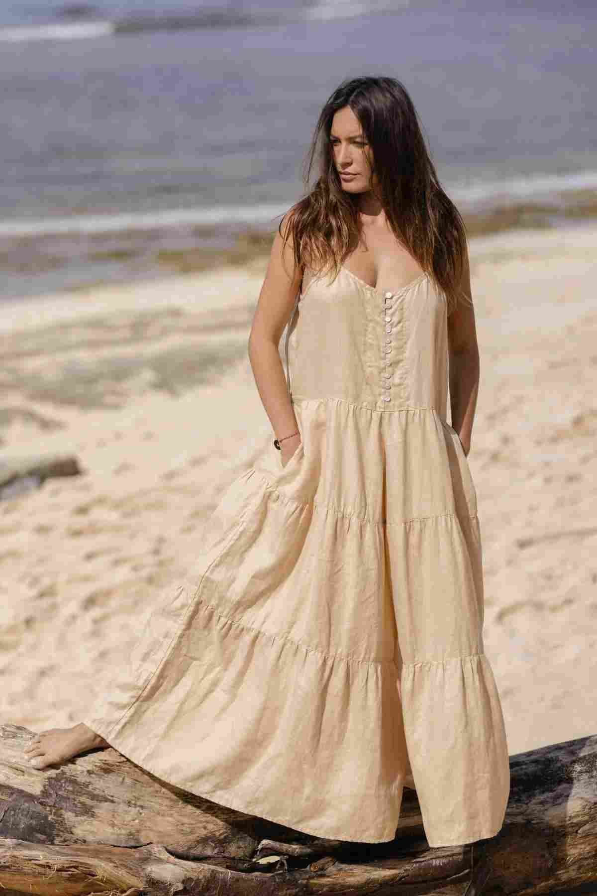 A woman stands on a sandy beach, wearing the La Mexicana Playsuit by Myrah Penaloza, crafted from 100% linen. The sea shimmers in the background as she gazes to the side with her hands resting on the adjustable straps. The sky is clear, and it's a sunny day.