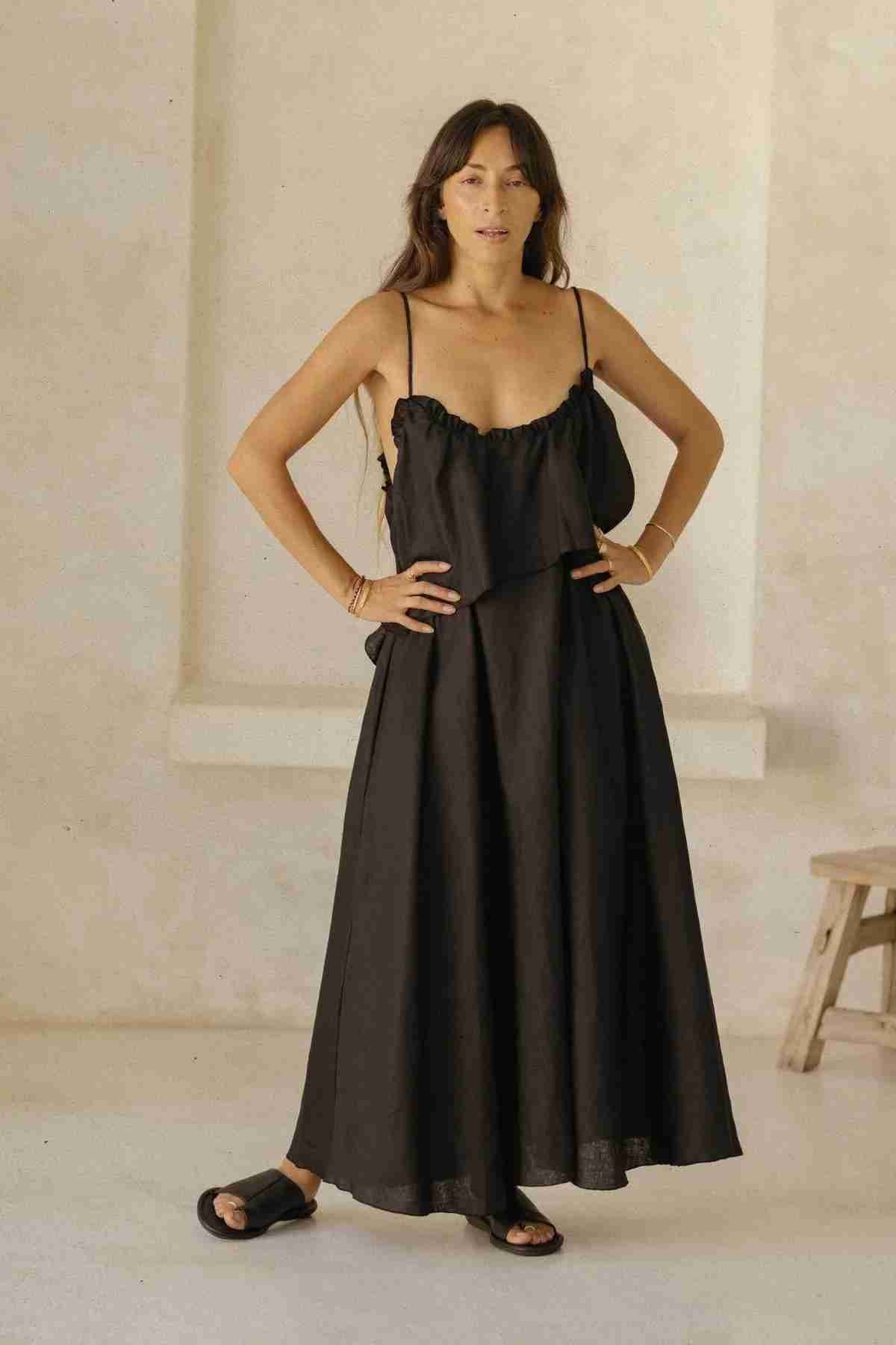 A person with long hair is standing with hands on hips, wearing the flowing black Linen Muse Gown by Myrah Penaloza and black slide sandals. The background features a neutral-toned wall and a wooden stool.