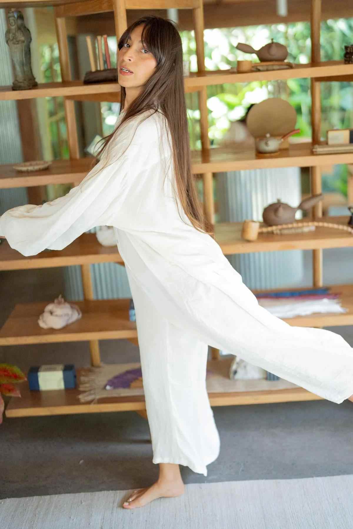 A person with long hair wearing a flowing white Kundalini Gown Playsuit Edition (Pre-Order) from Myrah Penaloza is standing on one leg in front of a wooden shelf filled with various items. The background shows greenery outside through a large window. The person appears to be balancing or performing a light dance move.
