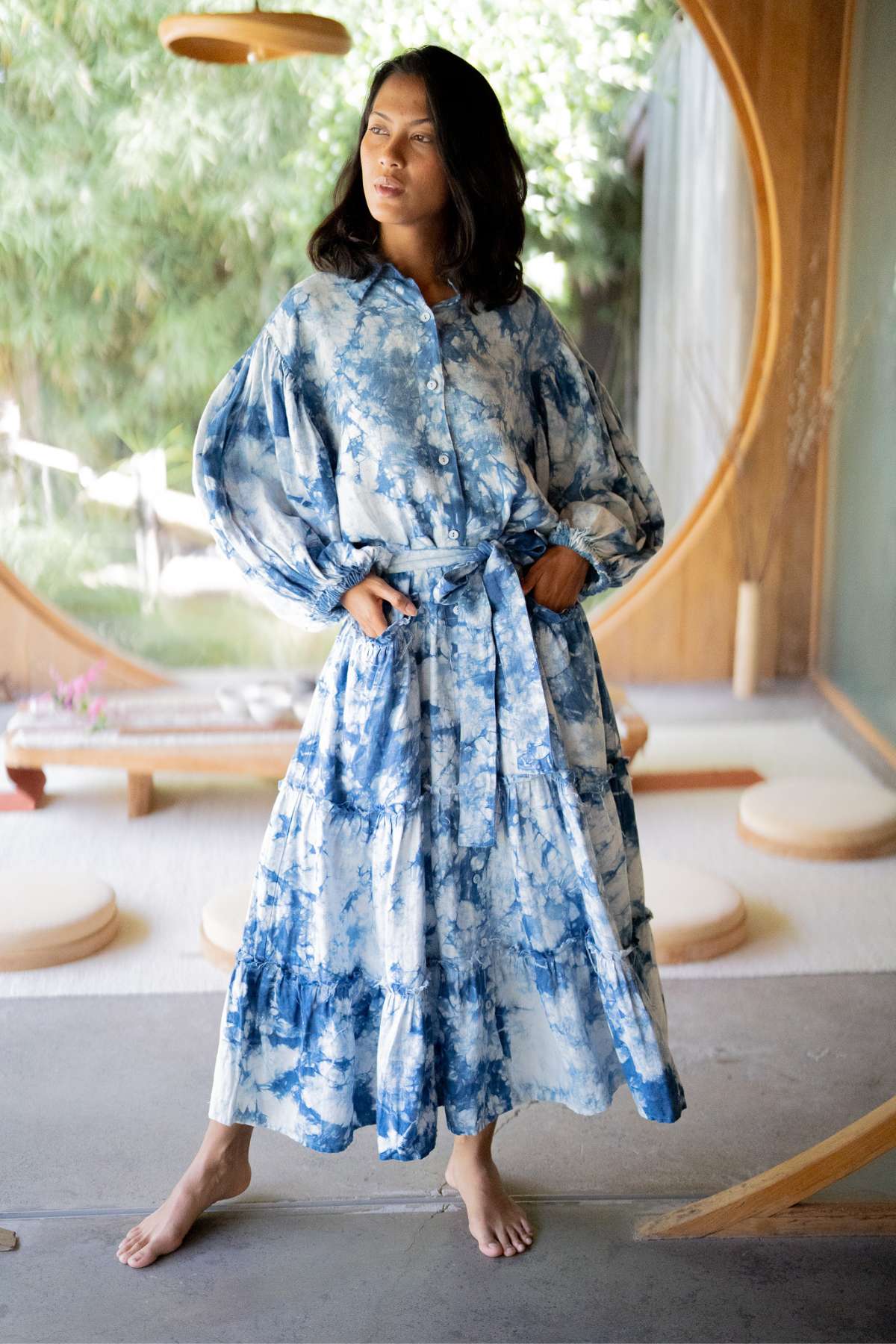 A person stands barefoot in a Myrah Penaloza La Dama Long Sleeves Ruffles dress, hand-dyed in indigo with a belt. They have long, dark hair and pose confidently in a room with a large circular window framing lush greenery outside.