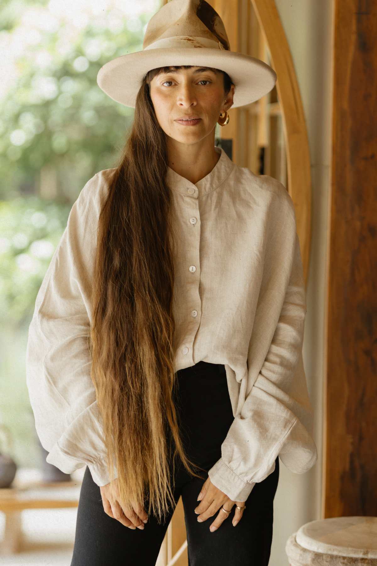 A person with long brown hair is standing indoors, wearing a wide-brimmed hat and the Cardenal Linen Top by Myrah Penaloza, paired with black pants. They are near a wooden door, with a softly blurred green background visible outside.