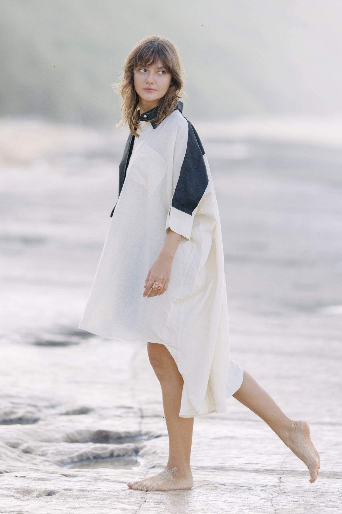 A barefoot woman, adorned in a versatile Linen Suka Button Down Long Top by Myrah Penaloza with black collar and cuffs, strolls along a sandy beach. The background is blurred, lending the scene a peaceful and serene ambiance. With shoulder-length hair drifting to the side, she subtly highlights the sustainable fabric of her attire.