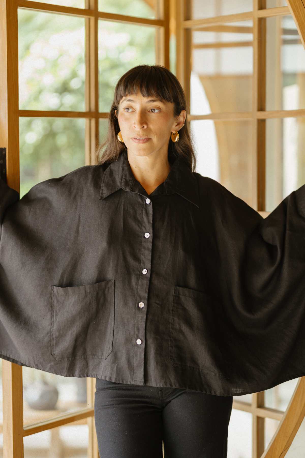 A person stands in a doorway wearing the Grace Linen Jacket by Myrah Penaloza, featuring large front pockets and wide sleeves. Their long hair with bangs complements the breathable design of the jacket as they gaze to the side. The background features wooden framing and a glimpse of greenery.