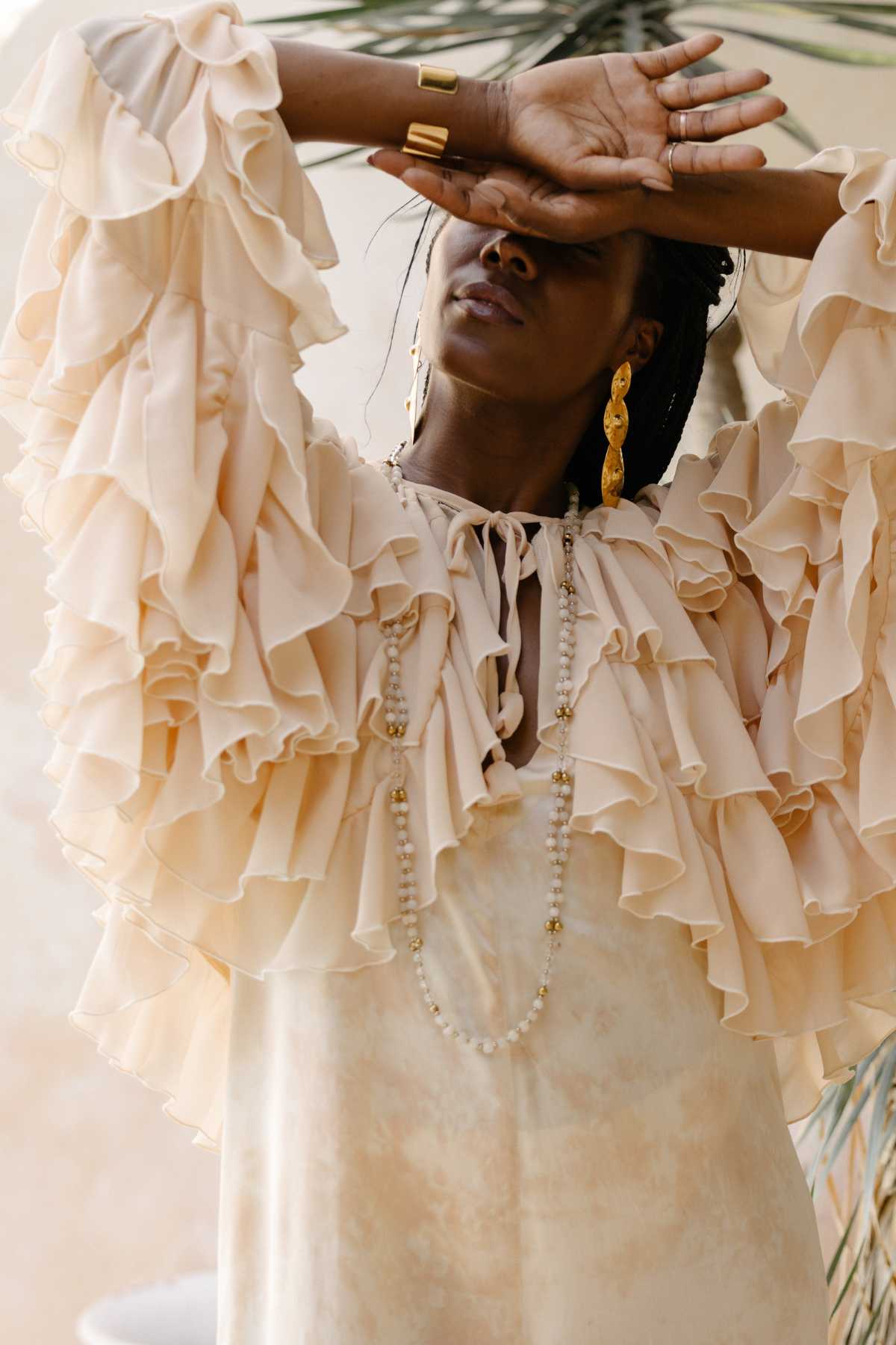 A person stands with one arm resting against their forehead, partially obscuring their face. They are adorned in the Anusara Bloom - Made To Order dress by Myrah Penaloza, a ruffled and flowy chiffon sheer silk piece in cream. Complementing the look are gold earrings and a long beaded necklace. This nature-inspired couture ensemble is set against a backdrop of blurred greenery and a light-colored wall.