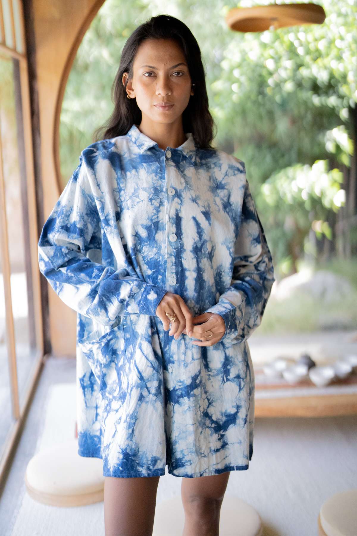 In a sunlit room, someone is wearing the Tender Jacket - Hand Tye Dyed Indigo from Myrah Penaloza. The background displays blurred greenery and round cushions on the floor, adding an eclectic touch to this cozy scene.