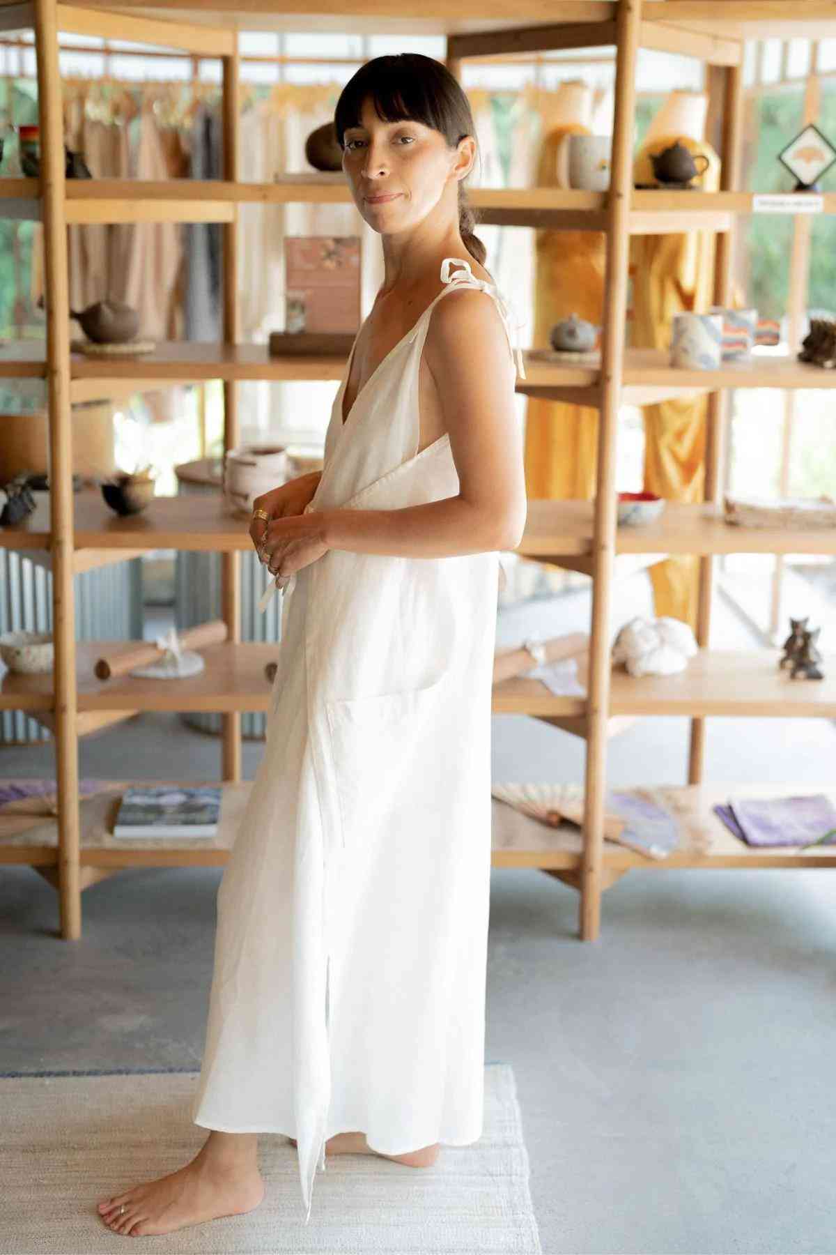 A person with dark hair and bangs is standing barefoot on a white rug in a minimalistic, naturally-lit room. They are wearing the Radiant Gown - Made To Order from Myrah Penaloza, a long, white sleeveless dress with ties at the shoulders. Wooden shelves filled with various items are in the background.