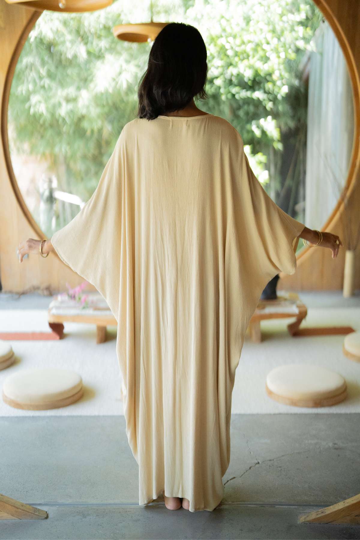 A person with long, dark hair stands barefoot, facing away in a flowing beige Virgo Moon robe by Myrah Penaloza. The robe's long sleeves and exquisite detailing harmonize with the circular architectural elements, while lush greenery is visible through large windows.