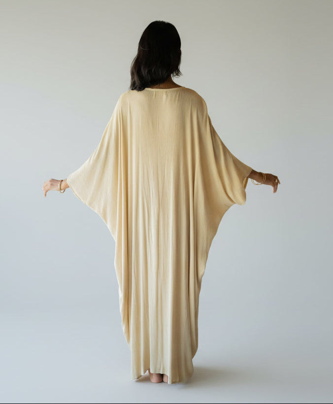 A person with long, dark hair stands barefoot, facing away in a flowing beige Virgo Moon robe by Myrah Penaloza. The robe's long sleeves and exquisite detailing harmonize with the circular architectural elements, while lush greenery is visible through large windows.