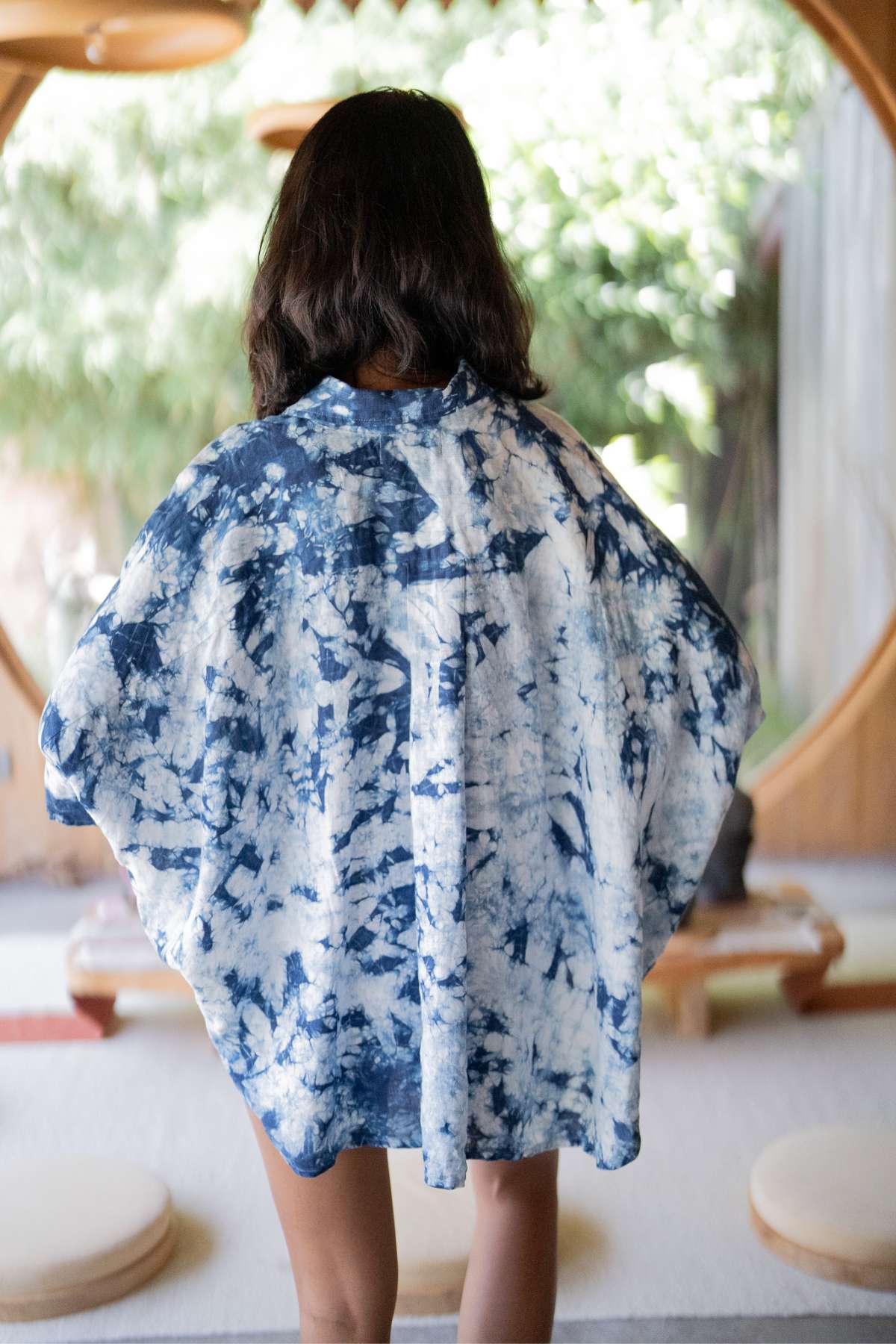 A person with dark hair stands facing away, wearing the Indigo Suka Set by Myrah Penaloza, made from 100% linen and ethically sourced materials. The background features a round doorway and a tranquil room bathed in soft lighting with elements of nature.