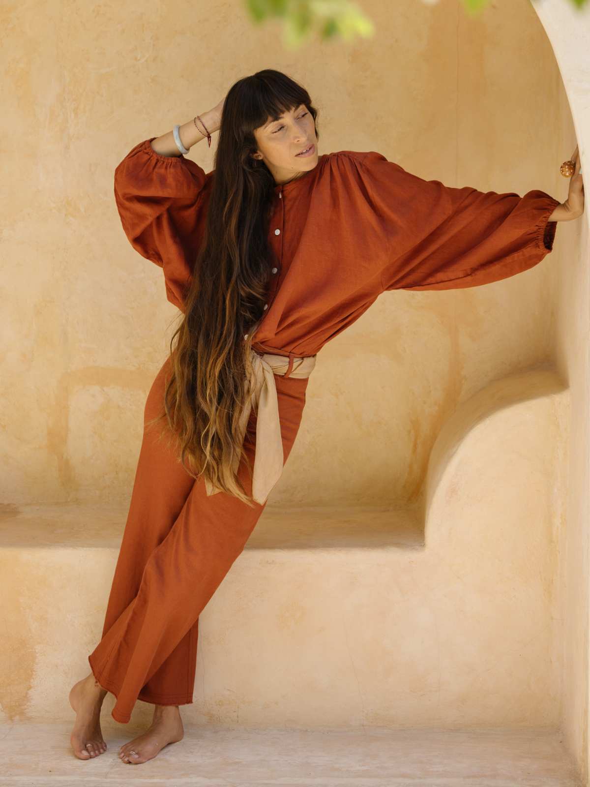 A woman with long hair and bangs leans against a beige stucco wall, wearing the Rice Linen Set by Myrah Penaloza. Her rust-colored blouse with billowy sleeves and matching rust-colored pants, crafted from elegant stone-washed linen, along with her barefoot pose create a relaxed and stylish atmosphere.