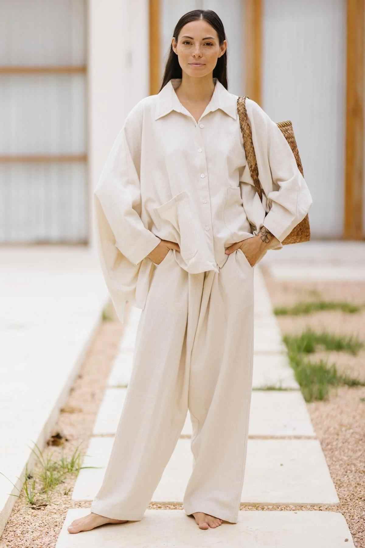 A person with long dark hair stands confidently in a light, flowing, oversized Grace Jacket & Mona Pant Set from Myrah Penaloza. They have their hands in their pockets and are carrying a woven shoulder bag. They are standing barefoot on a light stone pathway with a building in the background.