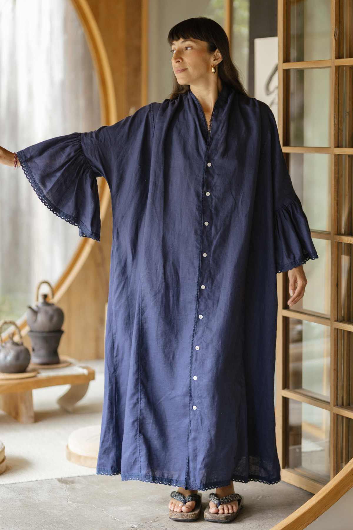 A person stands in a room wearing the Kyoto Duster by Myrah Penaloza, a long deep blue garment with flared sleeves that evokes ceremonial attire. They are near a wooden sliding door, and teapots rest on a low table in the background, creating a calm and cozy atmosphere.