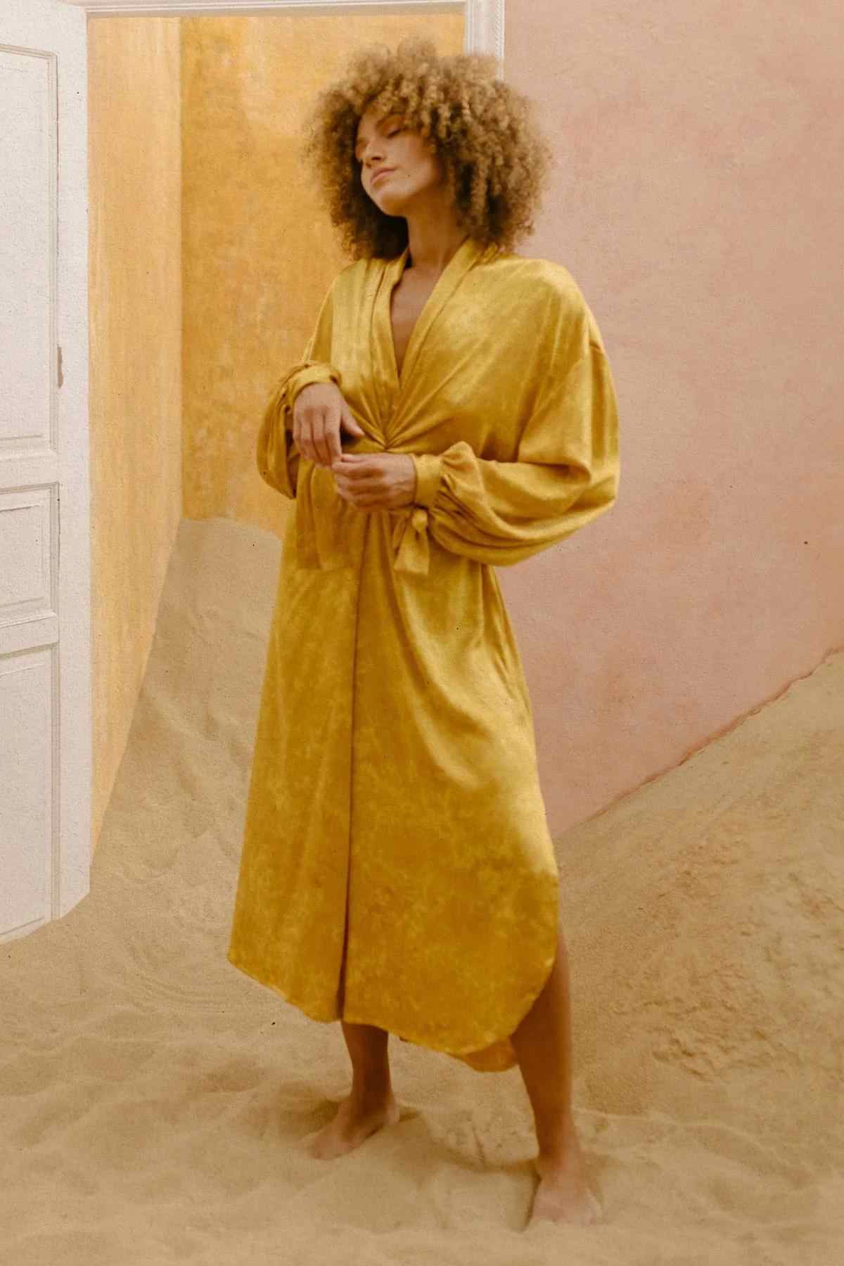 A person with curly hair stands barefoot in a room filled with sand, wearing the Silk Inanna Wrap Gown by Myrah Penaloza. The long, flowing, mustard-yellow dress features a tied waist and balloon sleeves. The room has a door on one side and walls painted in warm tones. The person has a serene expression.