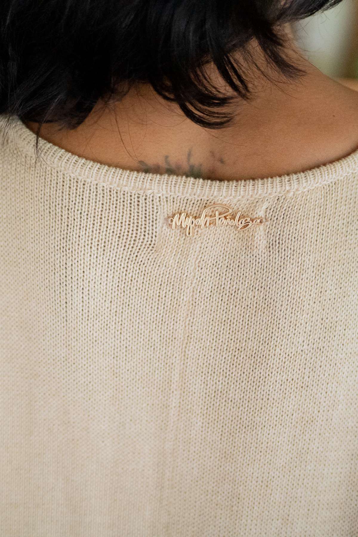 A close-up shows a person wearing the Henley Knitted Playsuit by Myrah Penaloza, featuring a beige cotton knit design. A small gold signature pin adorns the neckline, while a tattoo is partly visible on their neck. The individual's short, dark hair enhances their sophisticated yet casual style.