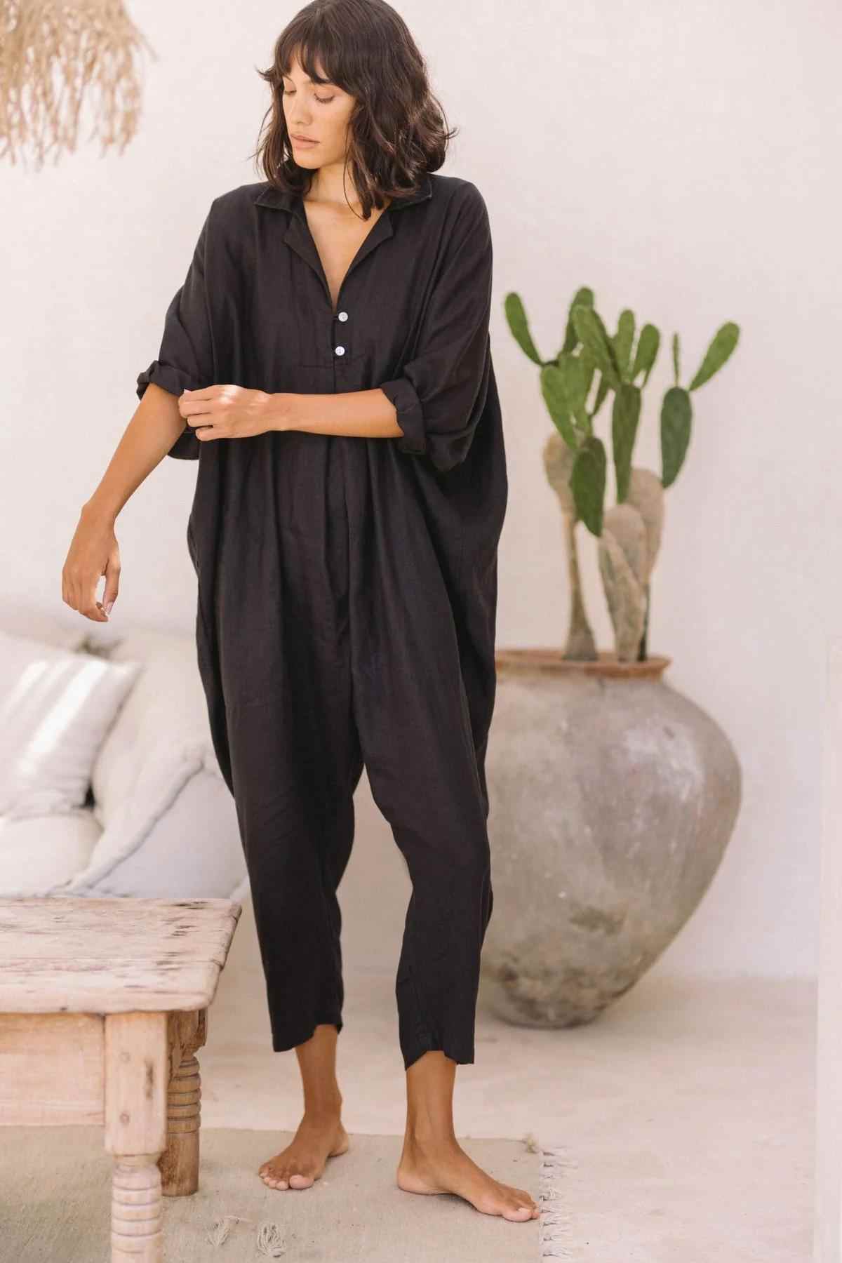 With shoulder-length dark hair, a person stands barefoot on a light-colored floor, wearing the Linen Sat Torri Playsuit by Myrah Penaloza. The loose-fitting black jumpsuit features wide dolman sleeves rolled up, which the person is adjusting. Behind them is a large potted cactus, a white wall, and a wooden table with cushions that exude retro 1980s inspired vibes.