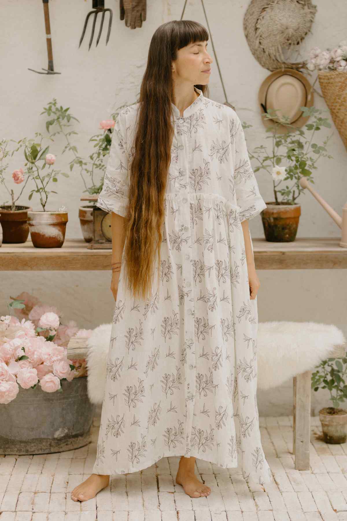 A woman stands barefoot in a garden-themed room, adorned in the long, white Bona Dress from the Cha Dao Tea Collection by Myrah Penaloza. The dress, made of 100% linen with delicate floral patterns and a limited edition Tea Leaf Eco Print, complements her very long hair worn loose. She gazes slightly to the side against a backdrop of potted plants, garden tools, and wicker baskets.