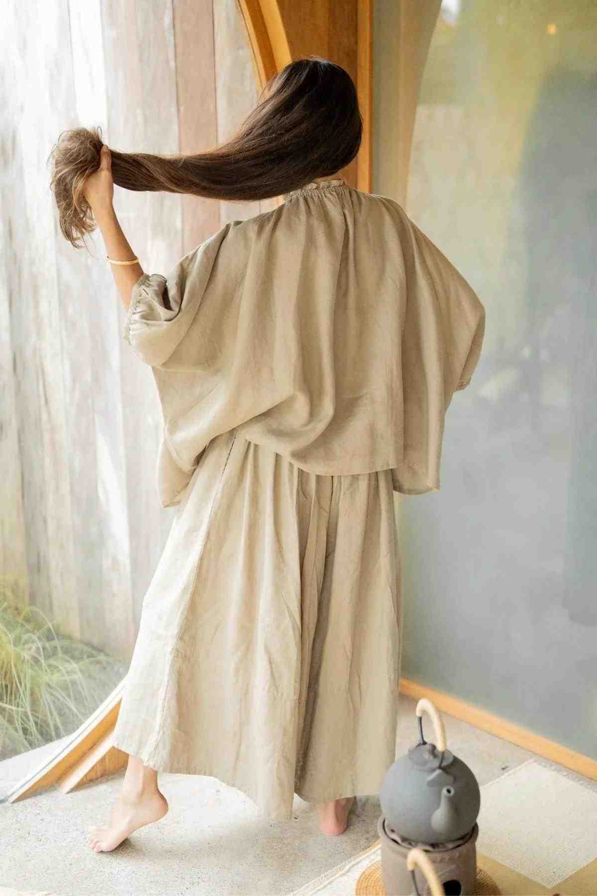 A person wearing a loose, light chocolate linen Playa Top & Pantalon Anco Set from Myrah Penaloza stands barefoot inside a room with wooden and glass elements. Their back is facing the camera, and they are holding up their long, dark hair with one hand, showcasing the flowing fabric of their outfit.