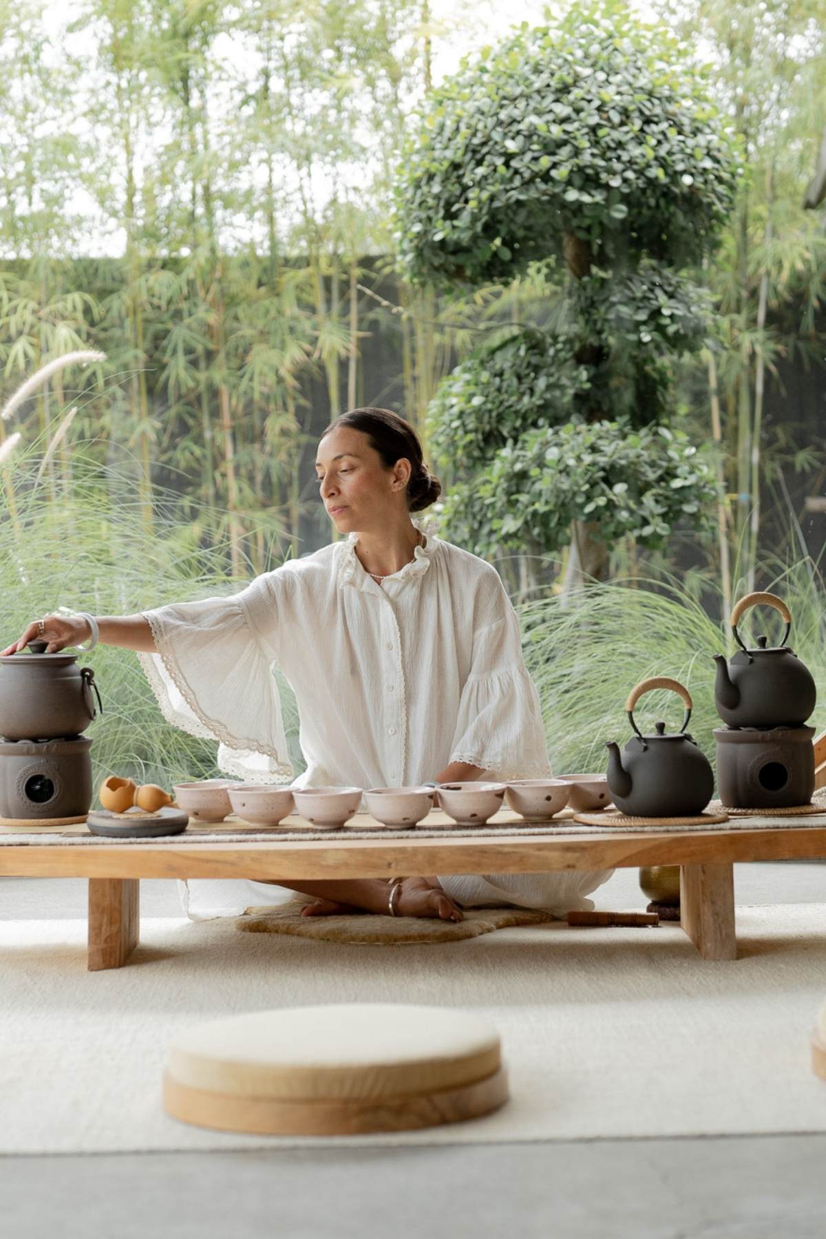 A person in a white dress embraces mindfulness through Myrah Penaloza’s "Embark on a Journey of Cha Dao: A way of Tea 1:1 Mentorship," preparing tea at a low wooden table in a serene outdoor setting. Ceramic teapots and cups are arranged on the table, surrounded by lush greenery that enhances the peaceful atmosphere.