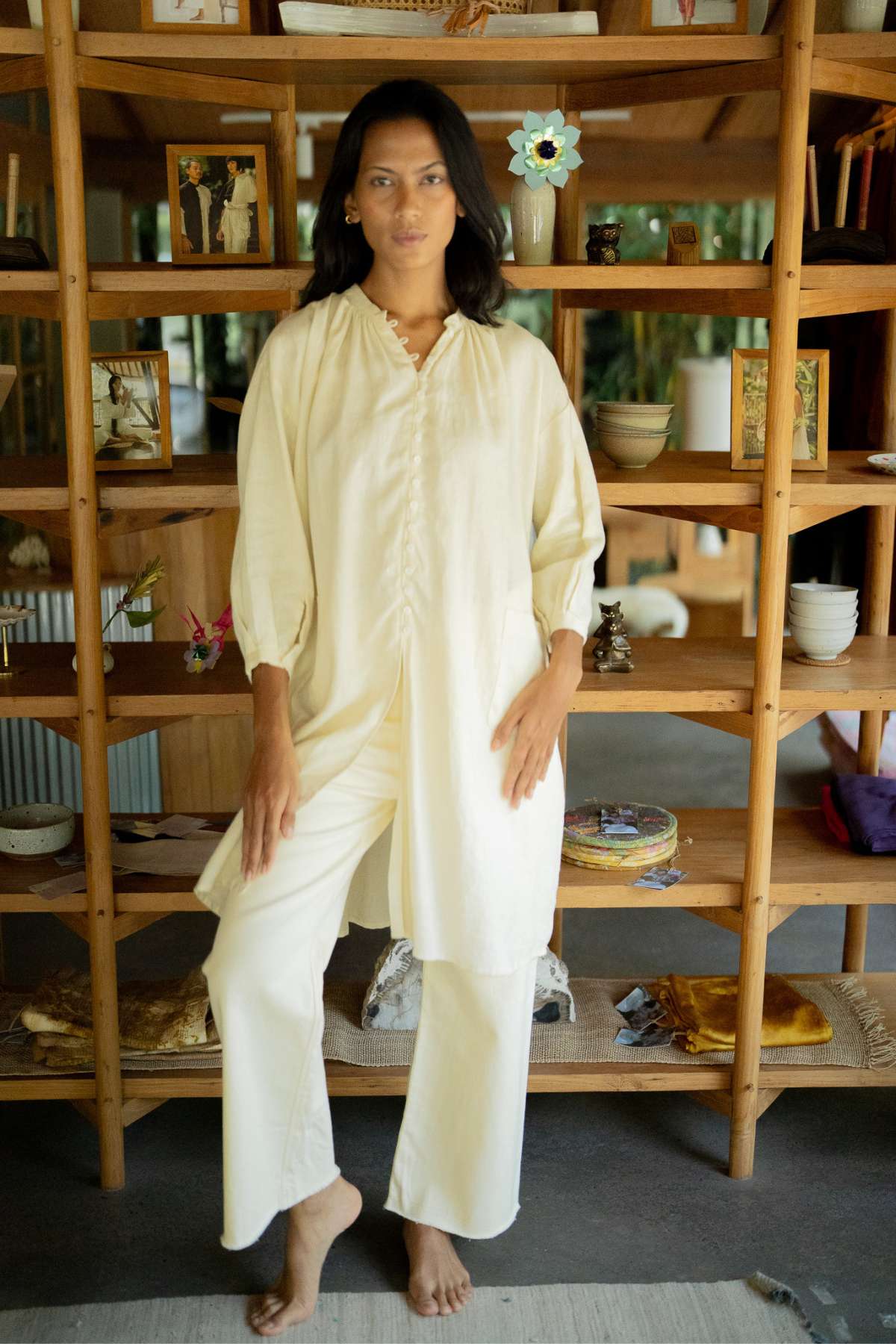 A woman wearing the Zazen Linen Top by Myrah Penaloza exudes effortless elegance as she stands barefoot in front of a wooden bookshelf filled with books, photos, and decorative items. Her long dark hair enhances the relaxed, casual indoor setting.