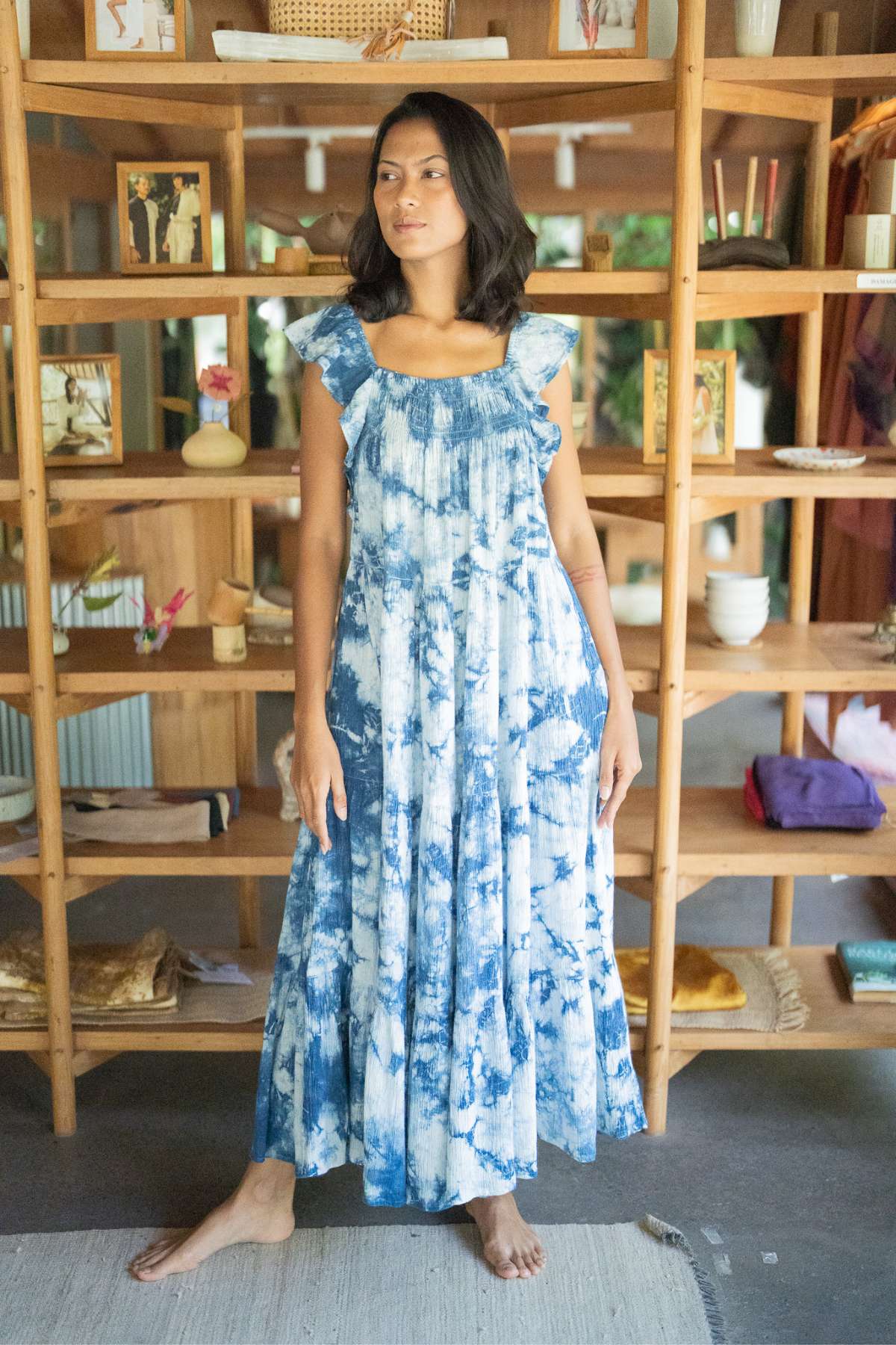 A woman wearing a sophisticated Myrah Penaloza Anna Yemaya Hand Tye Dyed Indigo - Black Friday stands barefoot on a rug in front of a wooden shelf filled with various items, including photos, vases, and decorative objects. She gazes to the side with a calm expression.