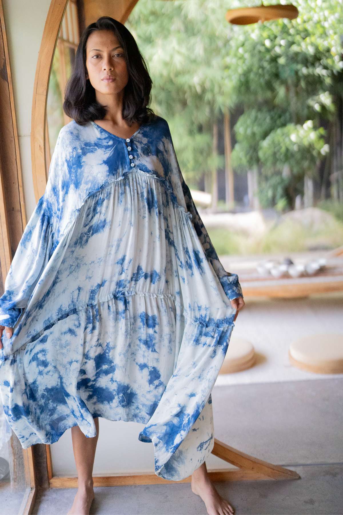 A person stands in a room with large windows, elegantly dressed in the Indigo Eva Gown by Myrah Penaloza, crafted from 100% silk. The gown's oversized design features stunning blue and white Indigo Dye patterns. The long sleeves enhance its elegance as greenery peeks through the panes, and circular cushions adorn the floor in this serene setting.