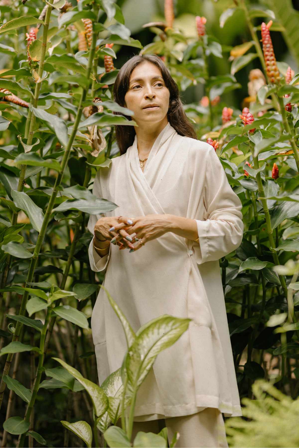 A person stands among tall, green plants and vibrant, red flowers. They are wearing an exquisite Guru Sheer Chiffon Top (Pre-Order) by Myrah Penaloza in off-white and have long, dark hair. The background is lush and full of greenery, suggesting a garden or natural setting.