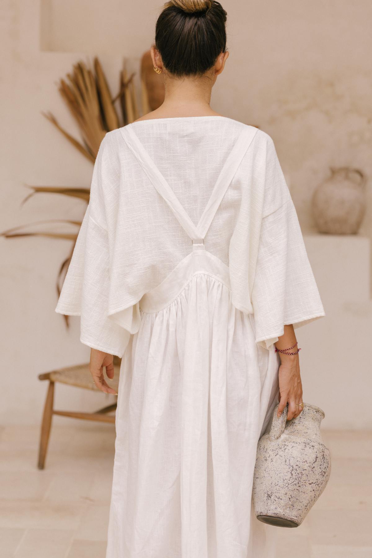 A person with their hair in a bun is seen from behind, wearing a flowing white May May Overall Skirt (Made to Order) by Myrah Penaloza featuring a V-shaped back design. They are holding a large, textured ceramic vase in their right hand. The background features neutral tones with dried plants and a rustic chair.