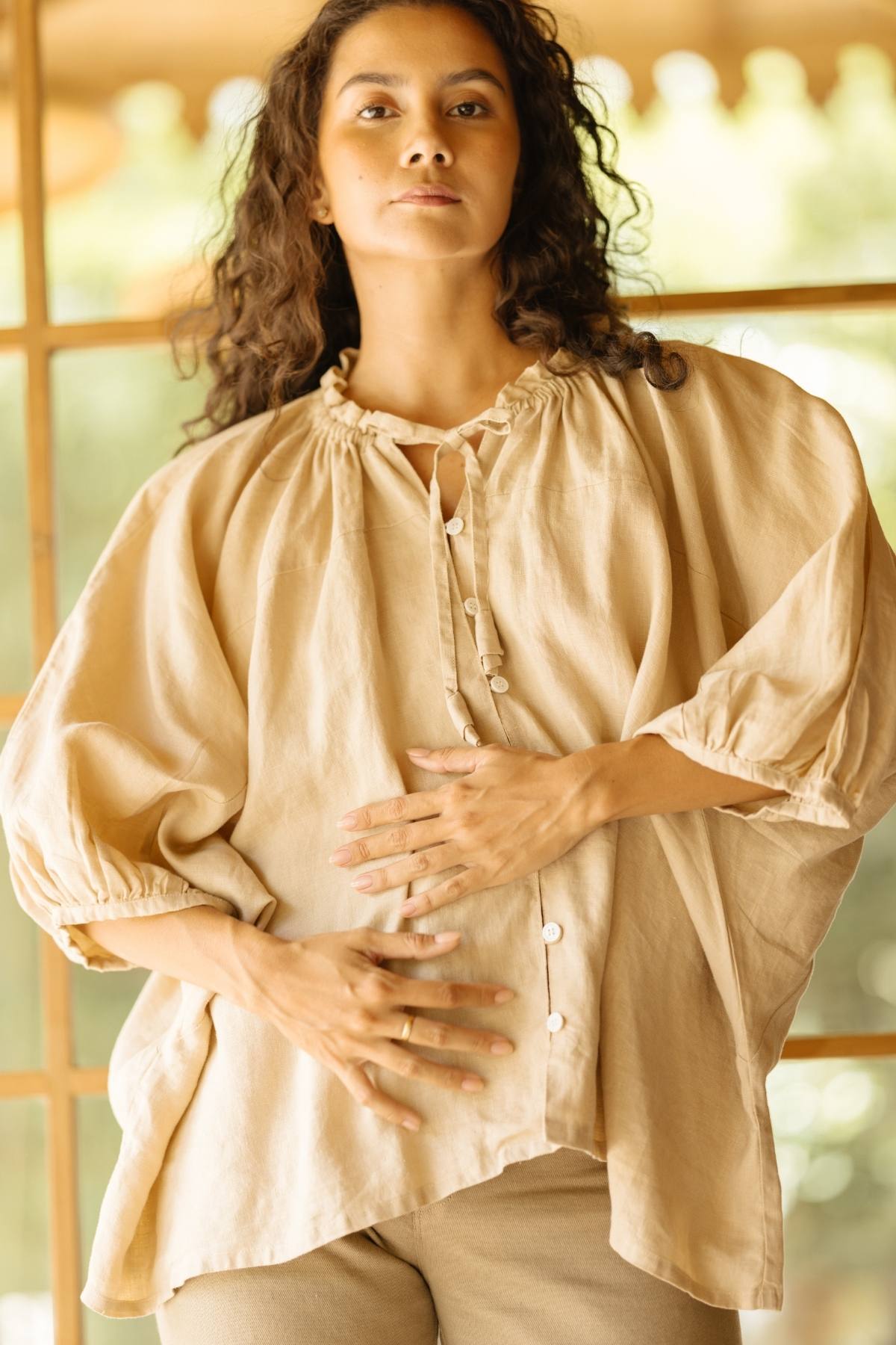 A person with long, wavy hair is wearing the Linen Playa Top from Myrah Penaloza. This loose-fitting, beige button-up blouse is made from 100% natural linen and features gathered details at the neckline and sleeves. They are standing indoors, gently placing their hands on their abdomen, with a wooden-framed window in the background.