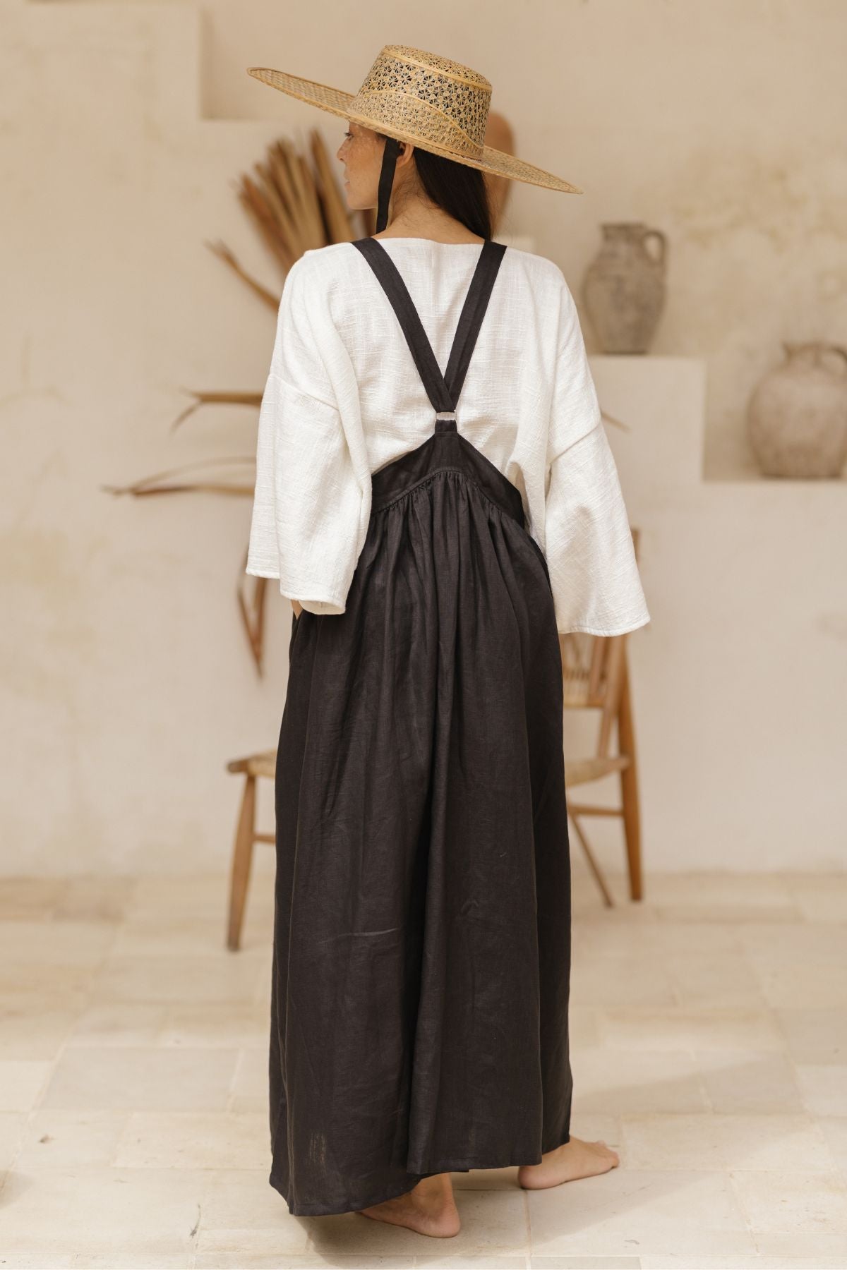 A person stands indoors on a light-colored floor, facing away from the camera. They are wearing a wide-brimmed straw hat, a loose white long-sleeved top, and the versatile **May May Overall Skirt (Made To Order)** by **Myrah Penaloza**. The background features a wooden chair and dried plants, enhancing the ambiance of a fairy garden.