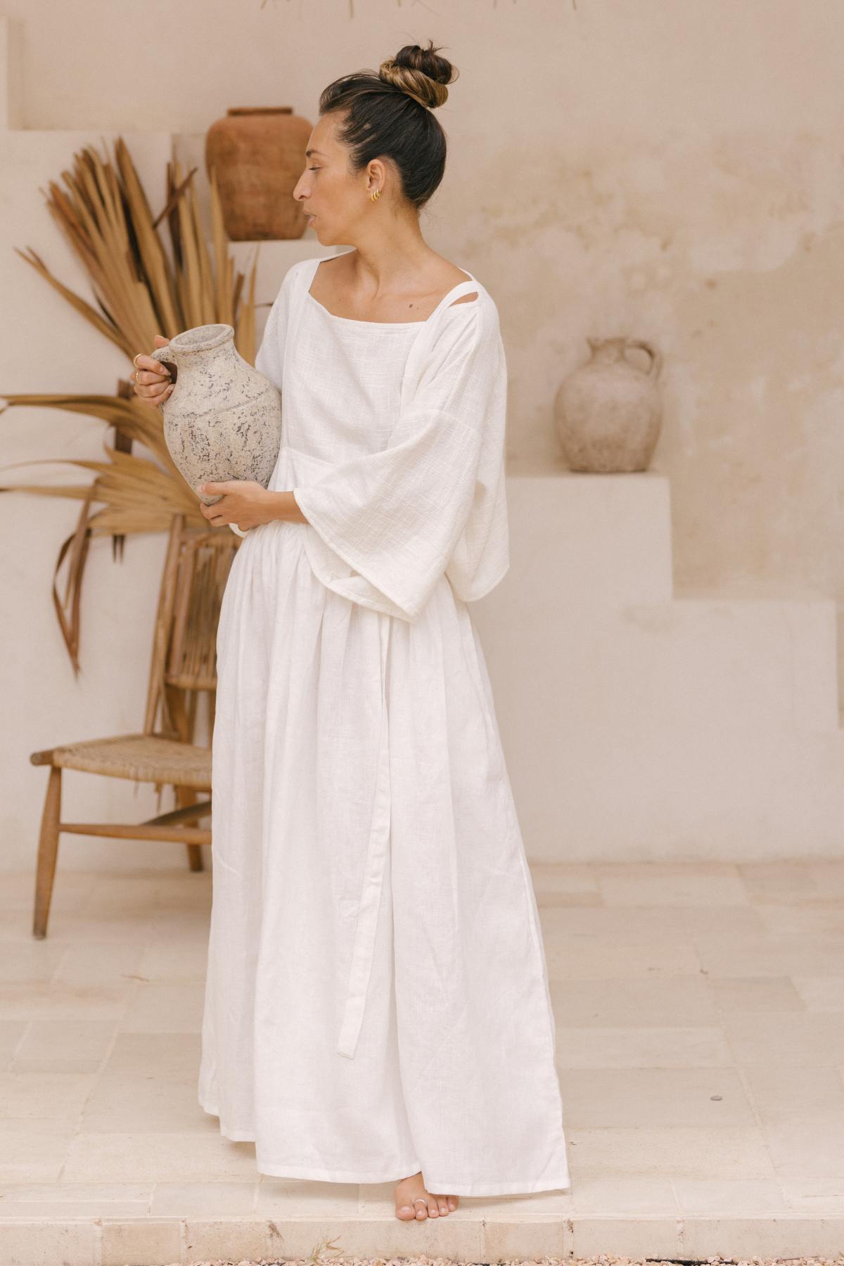 A woman in a May May Overall Skirt from Myrah Penaloza, made with 100% linen, is standing barefoot on a light-colored tiled floor, holding an aged clay jug. Her dark hair is pulled into a high bun. Behind her are dried palm leaves, a rustic chair, and a textured wall displaying a large pottery piece.