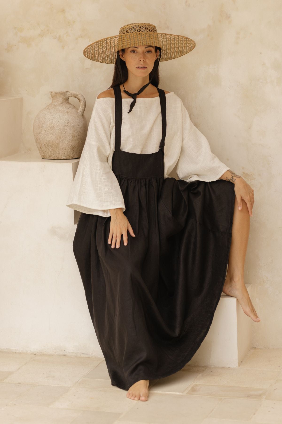 A woman sits on a white ledge, wearing a wide-brimmed straw hat, a white blouse with wide sleeves, and the chic May May Overall Skirt from Myrah Penaloza. One foot rests on the ledge while her other leg is bent, and she is barefoot. To her left, a rustic ceramic jug complements her serene setting.