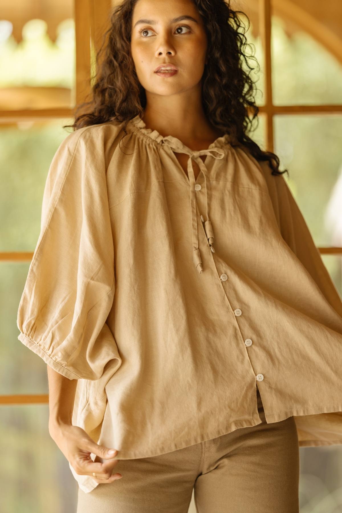 A woman with long curly hair wears the Linen Playa Top by Myrah Penaloza, a light chocolate linen blouse with buttons and balloon sleeves. She pairs it with beige pants and stands in front of a window with light streaming in, looking slightly to the side with a calm expression.