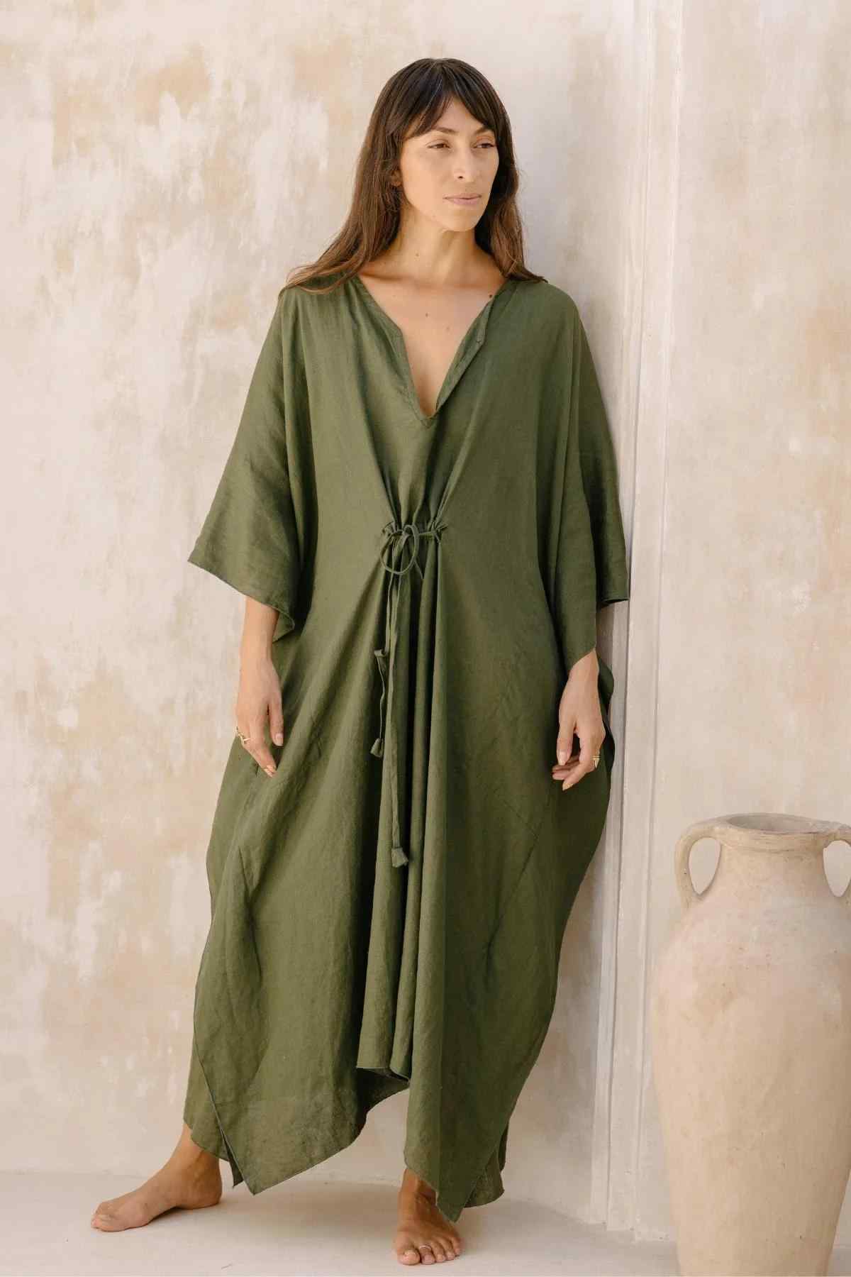 A woman stands against a textured beige wall, wearing a loose, flowing Myrah Penaloza Linen River Kaftan in olive green with a deep V-neckline and drawstring detail at the waist. The maternity-friendly dress complements her long, dark hair and calm, relaxed expression. A large, beige clay pot rests beside her.