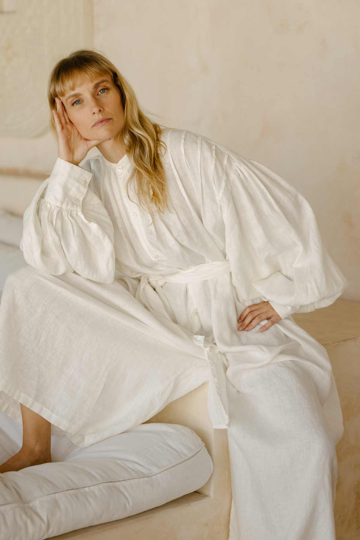 A person with long blond hair is sitting on a bed, slightly leaning, and wearing a flowing white Kundalini Gown Playsuit Edition made of 100% linen by Myrah Penaloza. They have a relaxed expression, resting their head on one hand. The background is softly lit with neutral tones.