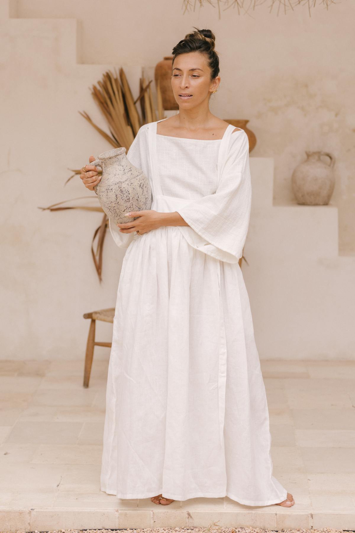 A person stands in a minimalist space, wearing a May May Overall Skirt from Myrah Penaloza. They hold a large, rustic jug with both hands. The background features a staircase, a wooden chair, and dried plants. The setting has a warm, natural tone.