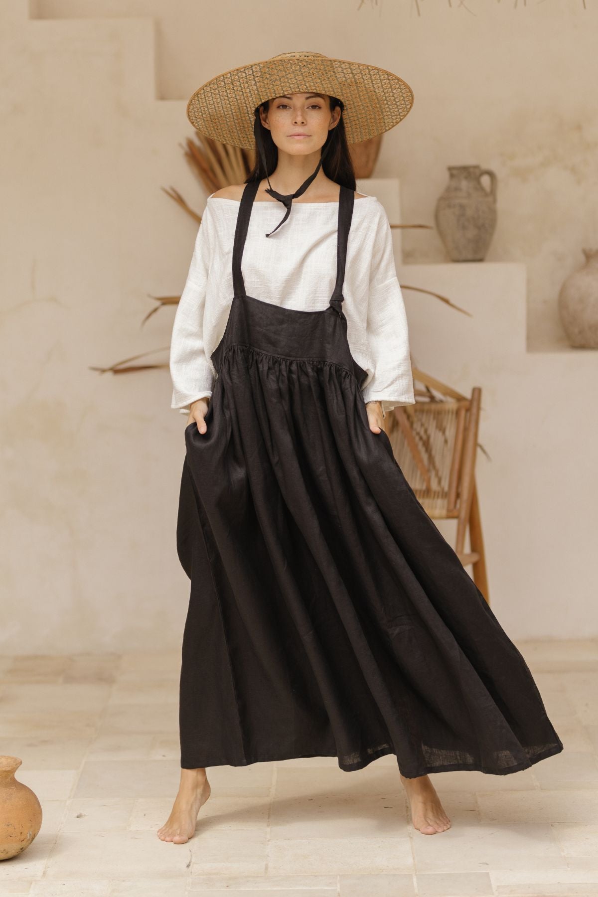 A person stands barefoot indoors, wearing a wide-brimmed straw hat and the Myrah Penaloza May May Overall Skirt (Made To Order) in long black crinkle linen over a white long-sleeve blouse. They pose with one leg slightly bent, creating movement in the skirt. The background includes pottery, a wicker chair, and decorative vases.