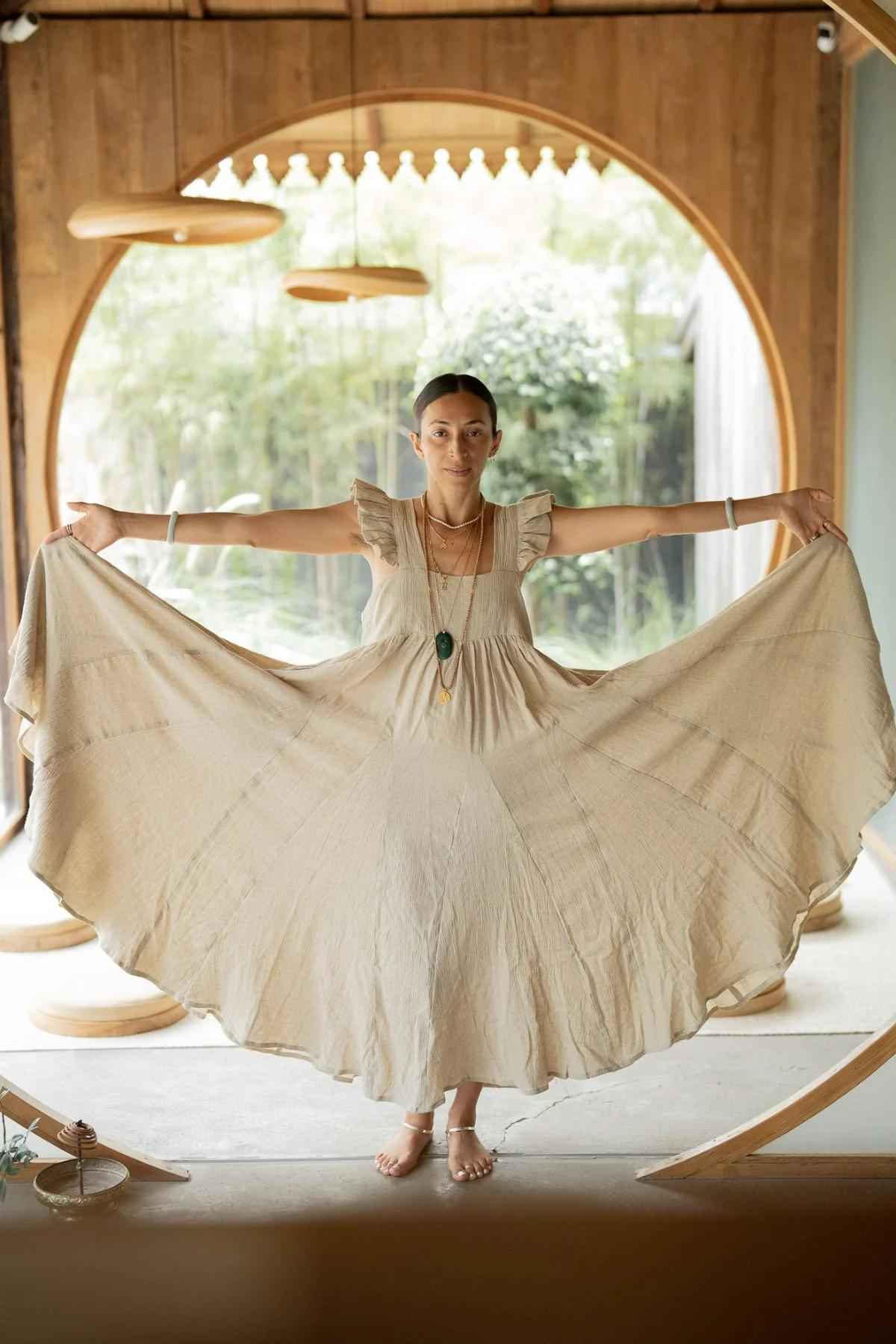 A woman stands indoors, holding out the sides of her long, beige Linen Anna Yemaya Gown by Myrah Penaloza to display its full width. She is barefoot and wears several necklaces. The background features a large, round window with a view of greenery outside. The room has wooden elements.