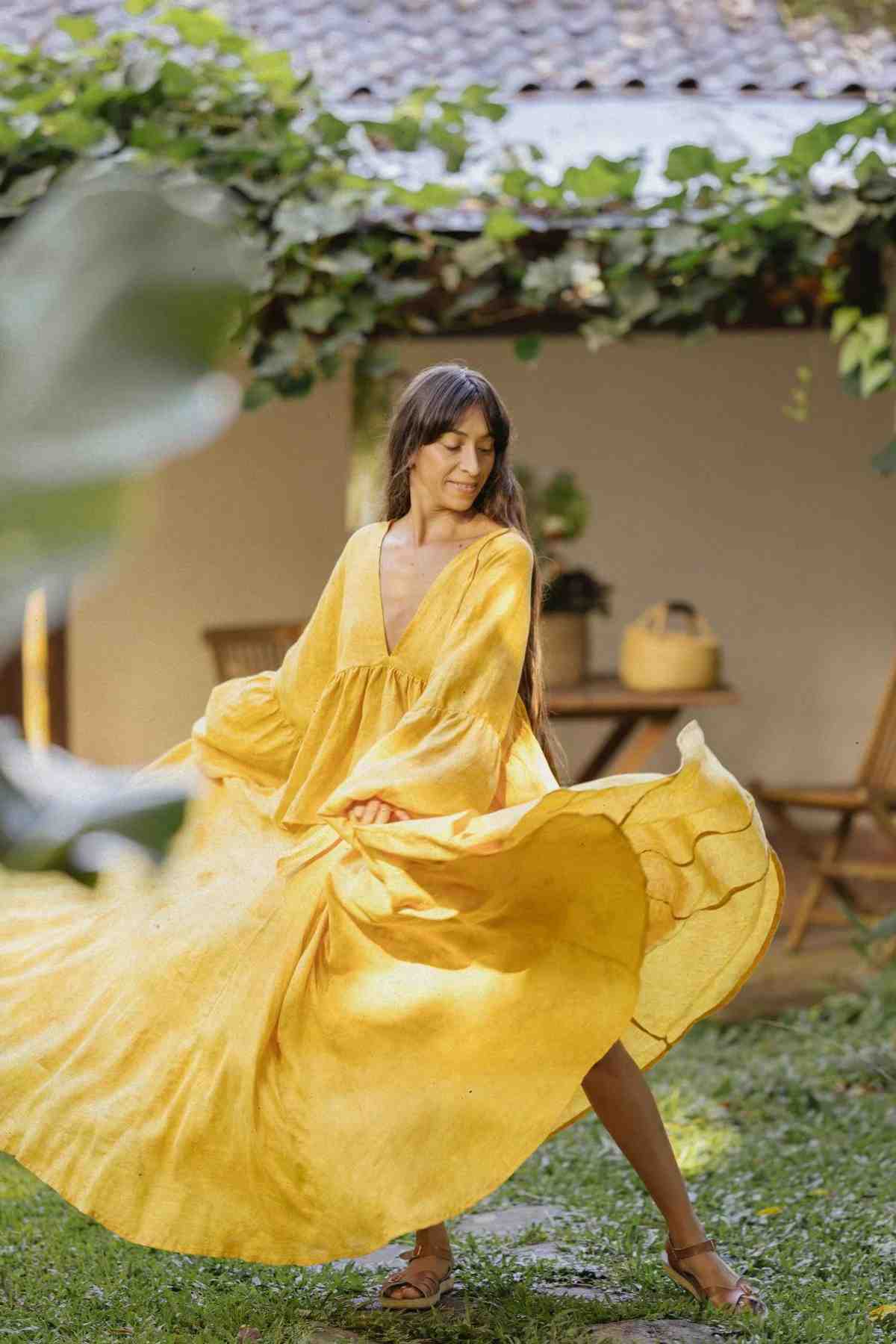 A woman with long hair twirls in a flowy, yellow Linen Eva Top & Orisa Dance Skirt Set by Myrah Penaloza, in an outdoor garden setting. She is surrounded by greenery, including a covered walkway and ivy, with a wooden table and chairs in the background.