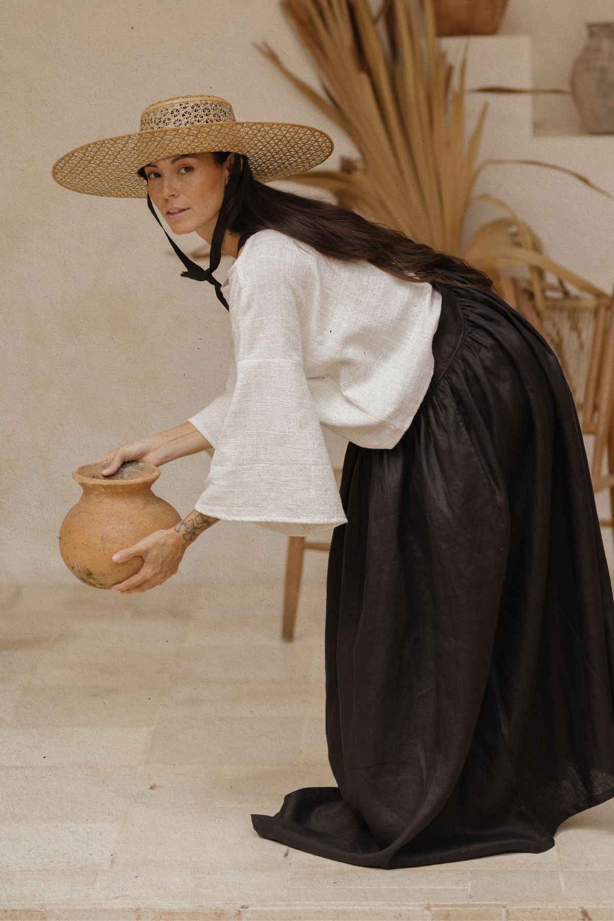 A woman, adorned in a wide-brimmed straw hat, white long-sleeve blouse, and the exquisite May May Overall Skirt from Myrah Penaloza's made-to-order collection, bends down slightly while holding a large clay pot. The background features dried plants and a neutral-toned wall, evoking a rustic, earthy ambiance.