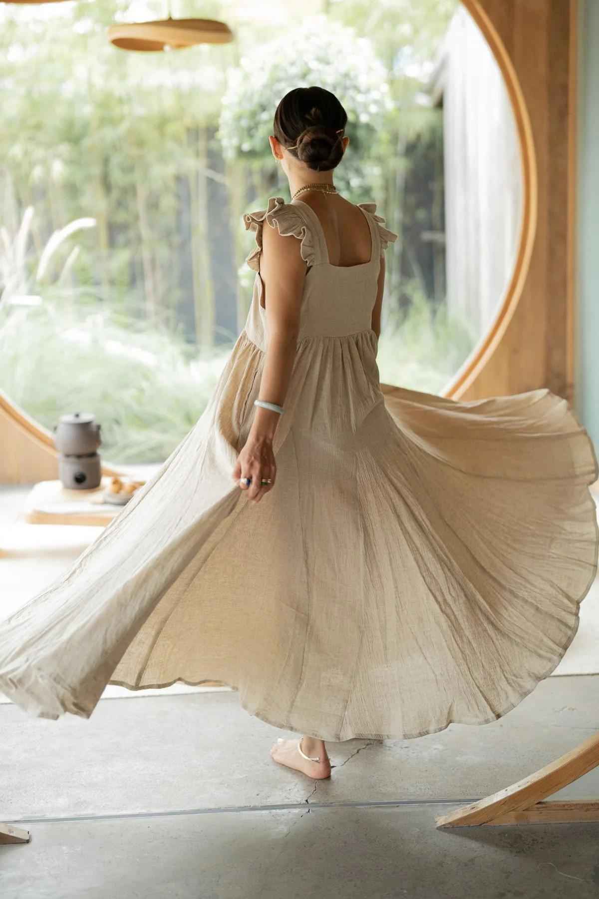 A woman twirls gracefully in a flowing Linen Anna Yemaya Gown by Myrah Penaloza, with her back to the camera. She is standing in a room with a large circular window, through which lush greenery is visible. The setting appears serene and natural, with soft lighting and minimalistic decor.