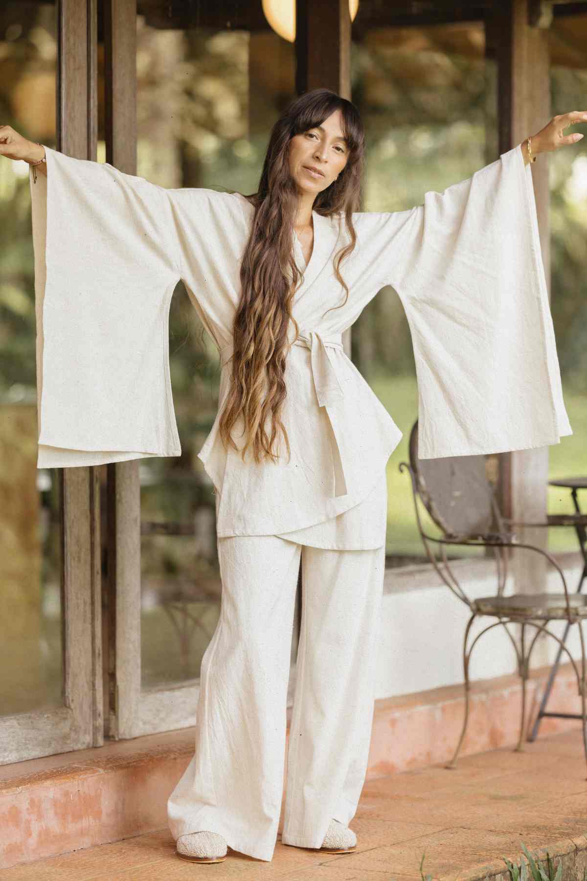 A woman with long brown hair is standing outside in front of a glass window and patio with her arms outstretched. She is wearing the Dewi Set (3 of a kind) from Myrah Penaloza, a light-colored, loose-fitting outfit made of hypoallergenic linen cotton crepe, featuring wide sleeves and wide-legged pants. A round table and chairs are partially visible in the background.