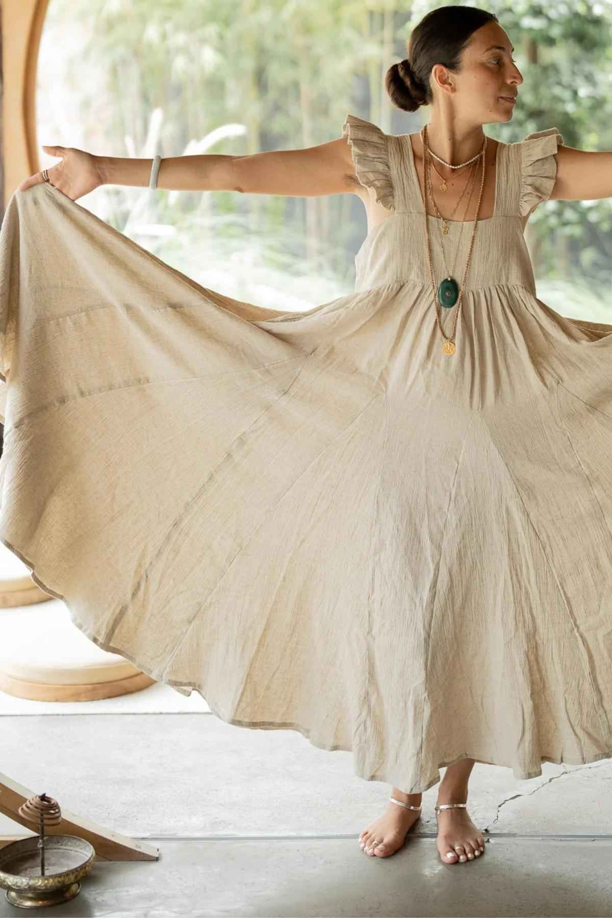 A person stands indoors with outstretched arms, displaying a flowing Linen Anna Yemaya Gown by Myrah Penaloza. They have their hair tied back and are wearing necklaces and sandals. The background includes wooden elements and greenery outside the window.