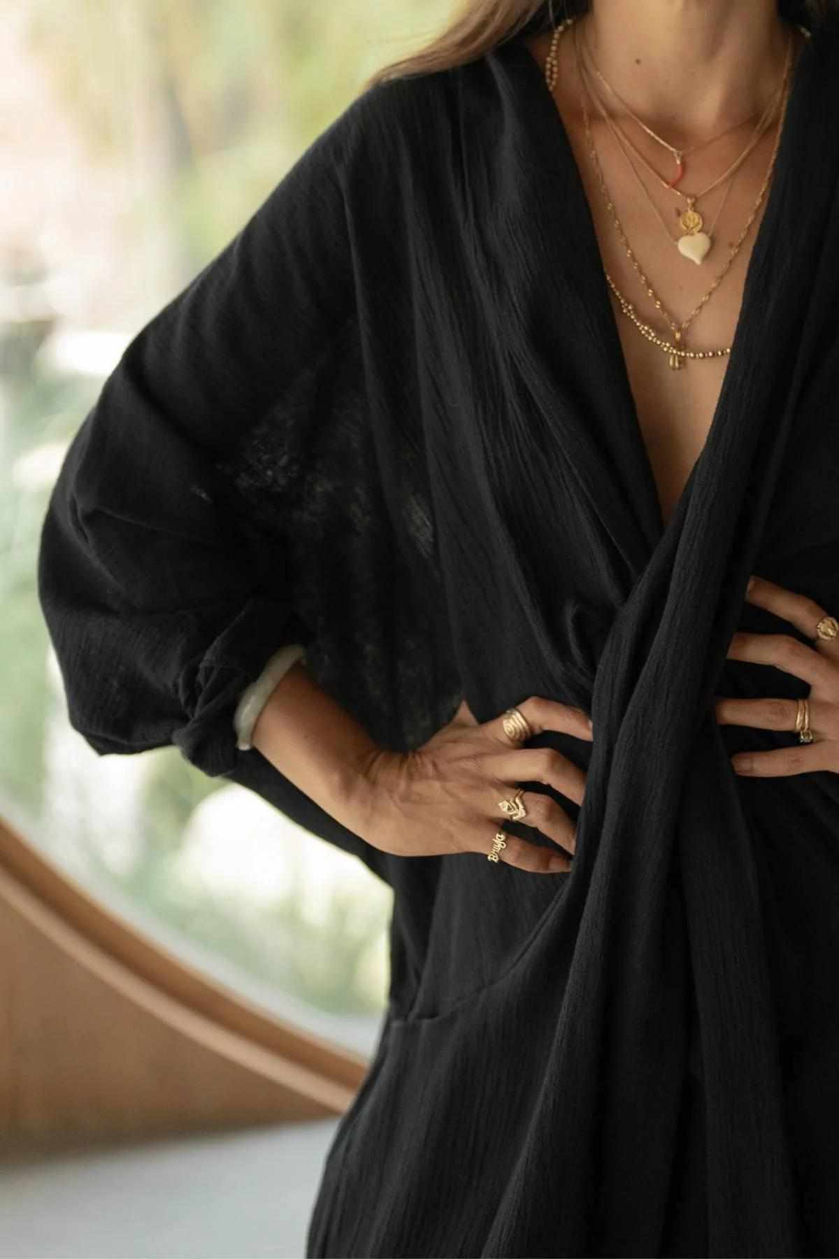 An individual wearing a black, long-sleeved blouse with a deep V-neckline is shown from the neck to the waist. They adorn multiple gold necklaces and rings. The background is softly blurred, accentuating a circular window and some greenery, infusing an air of elegance reminiscent of the Crinkle Linen Amma Playsuit by Myrah Penaloza.