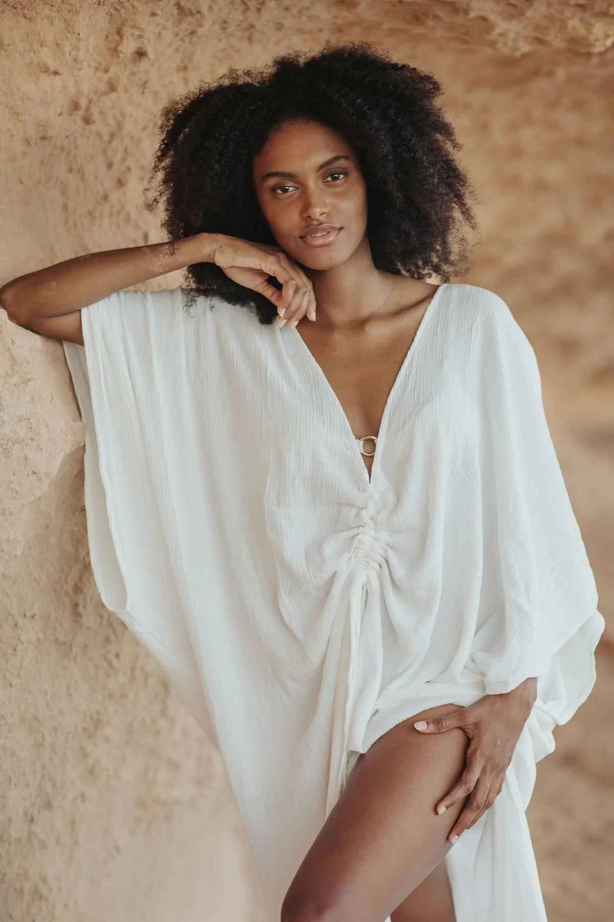 A woman with voluminous curly hair is leaning against a textured wall, elegantly dressed in Myrah Penaloza's Limited Edition Virgo Kaftan made of luxe bamboo rayon. With its deep V-neck and cinched waist, the kaftan exudes sophisticated fashion. She gazes at the camera with a serene expression, one leg slightly bent and her hand resting on her thigh.