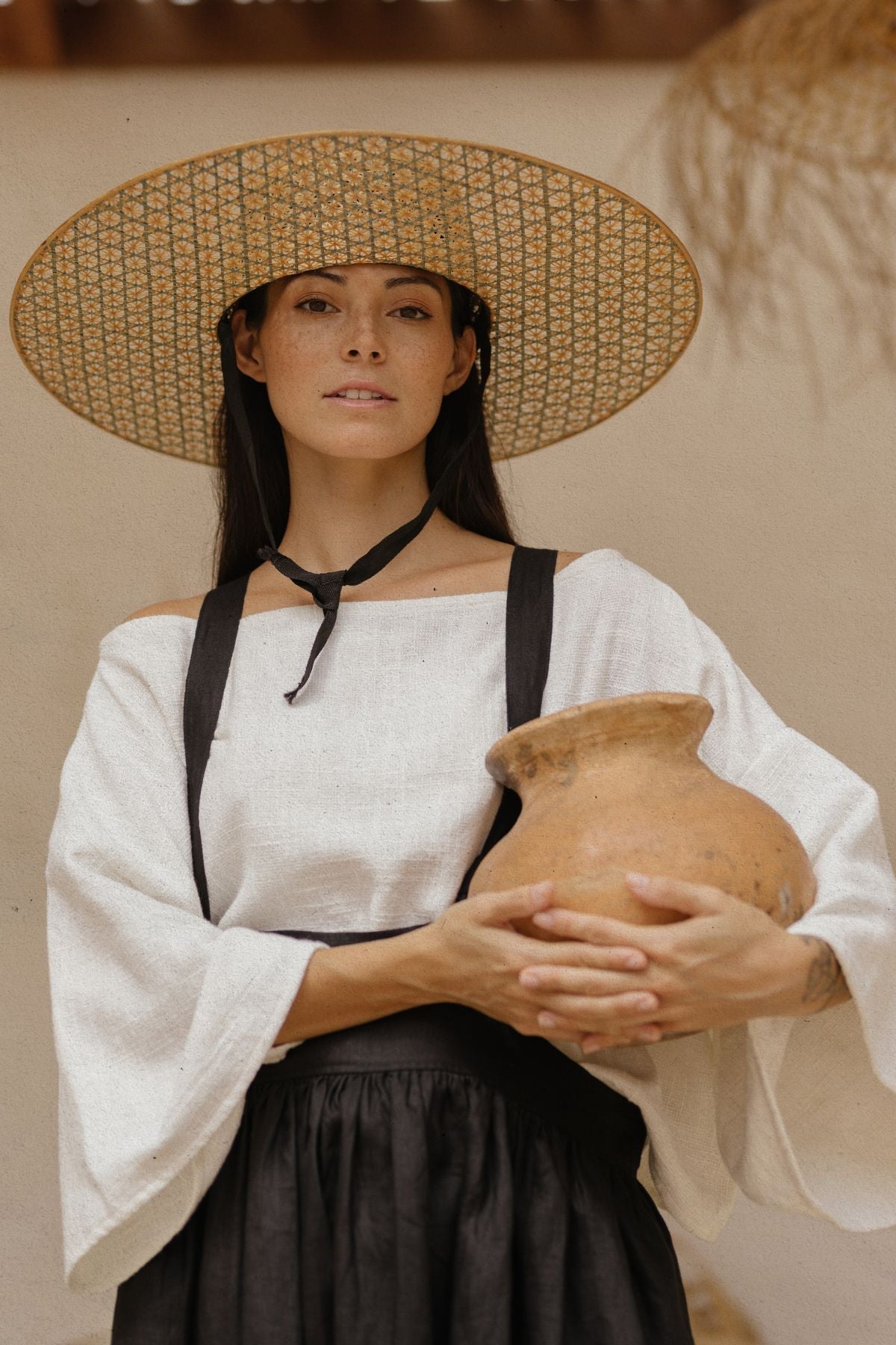 A person with long dark hair, wearing a wide-brimmed hat, a white blouse, and the May May Overall Skirt by Myrah Penaloza, is holding a large brown clay pot. The person stands against a plain background and looks at the camera with a neutral expression.