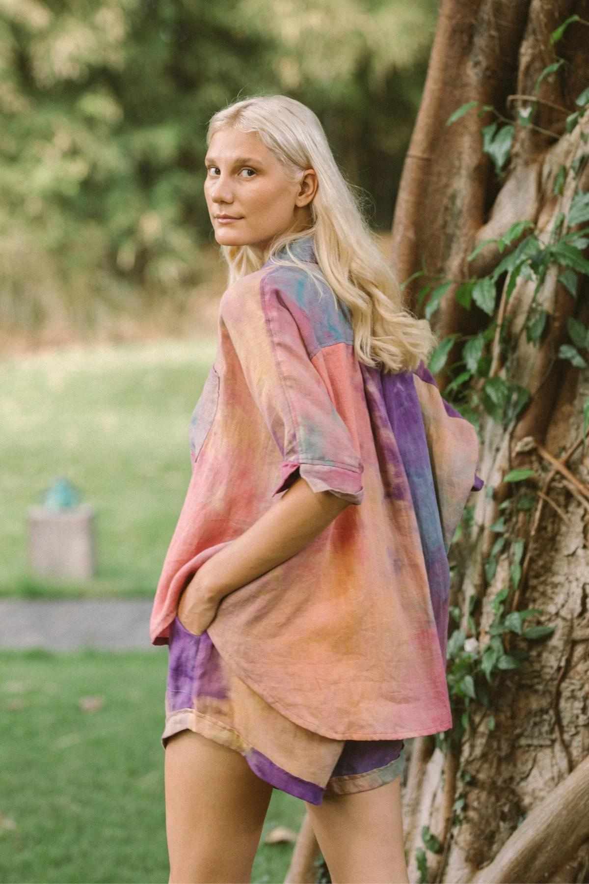 A person with long blonde hair stands outdoors in front of a large tree, wearing the vibrant and colorful Rainbeau Linen Suka Set by Myrah Penaloza. They glance over their shoulder towards the camera, surrounded by lush greenery.